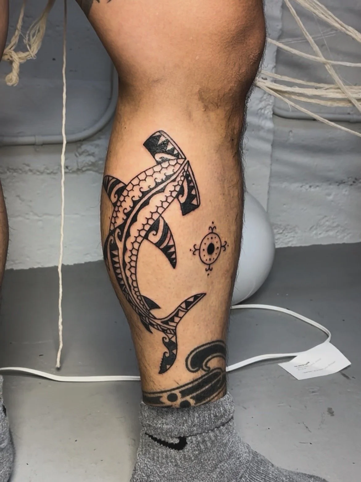 Tattoo of a black tribal-style shark on a person's thigh, with a small circular design nearby.