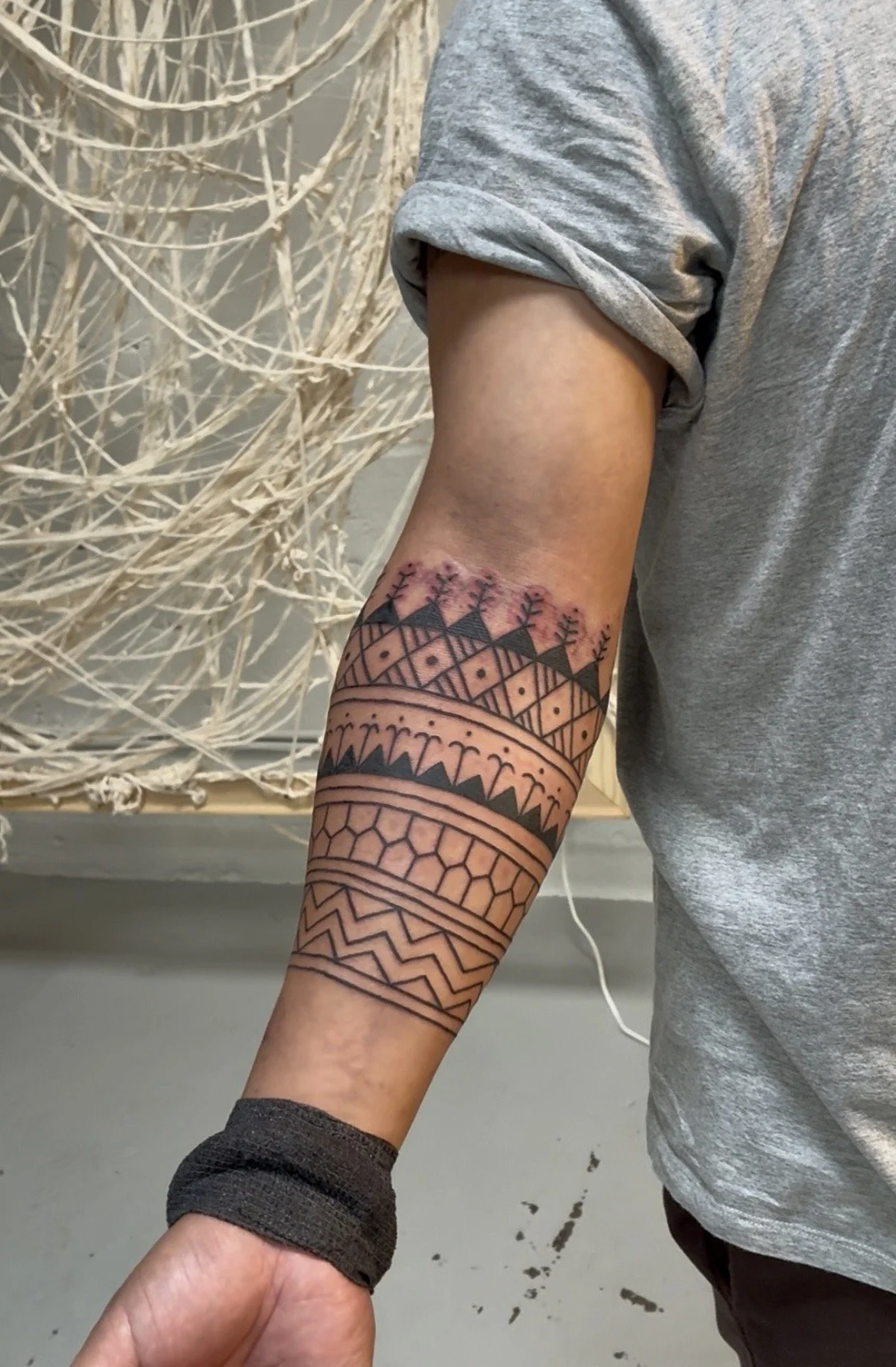 Person with a geometric patterned tattoo on their arm, wearing a gray rolled-up sleeve shirt, standing next to a artistic white wire sculpture.
