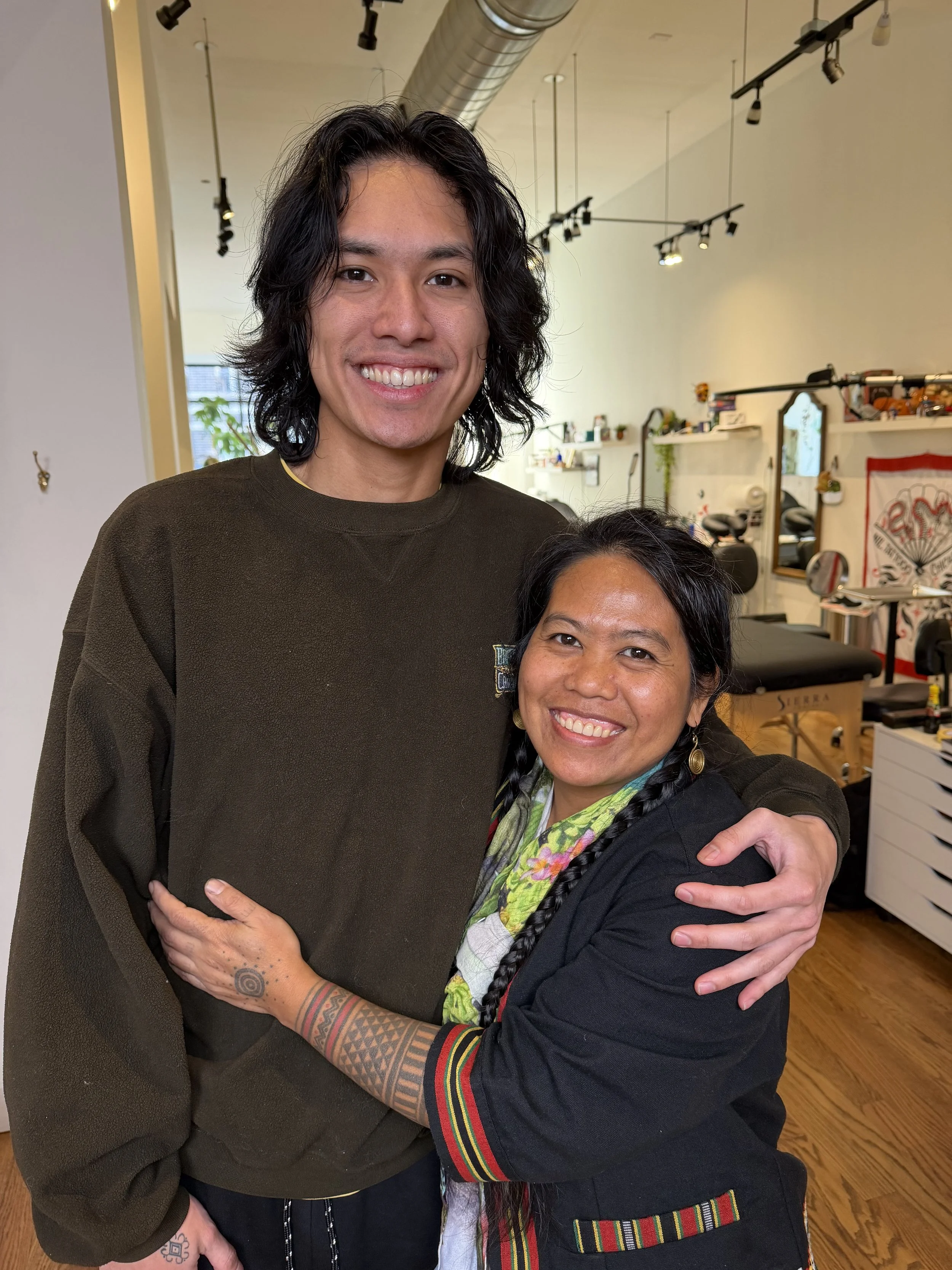 Two people hugging indoors, with visible tattoos and jewelry, smiling at the camera. The background shows a studio or creative space with mirrors, shelves, plants, and ceiling track lighting.