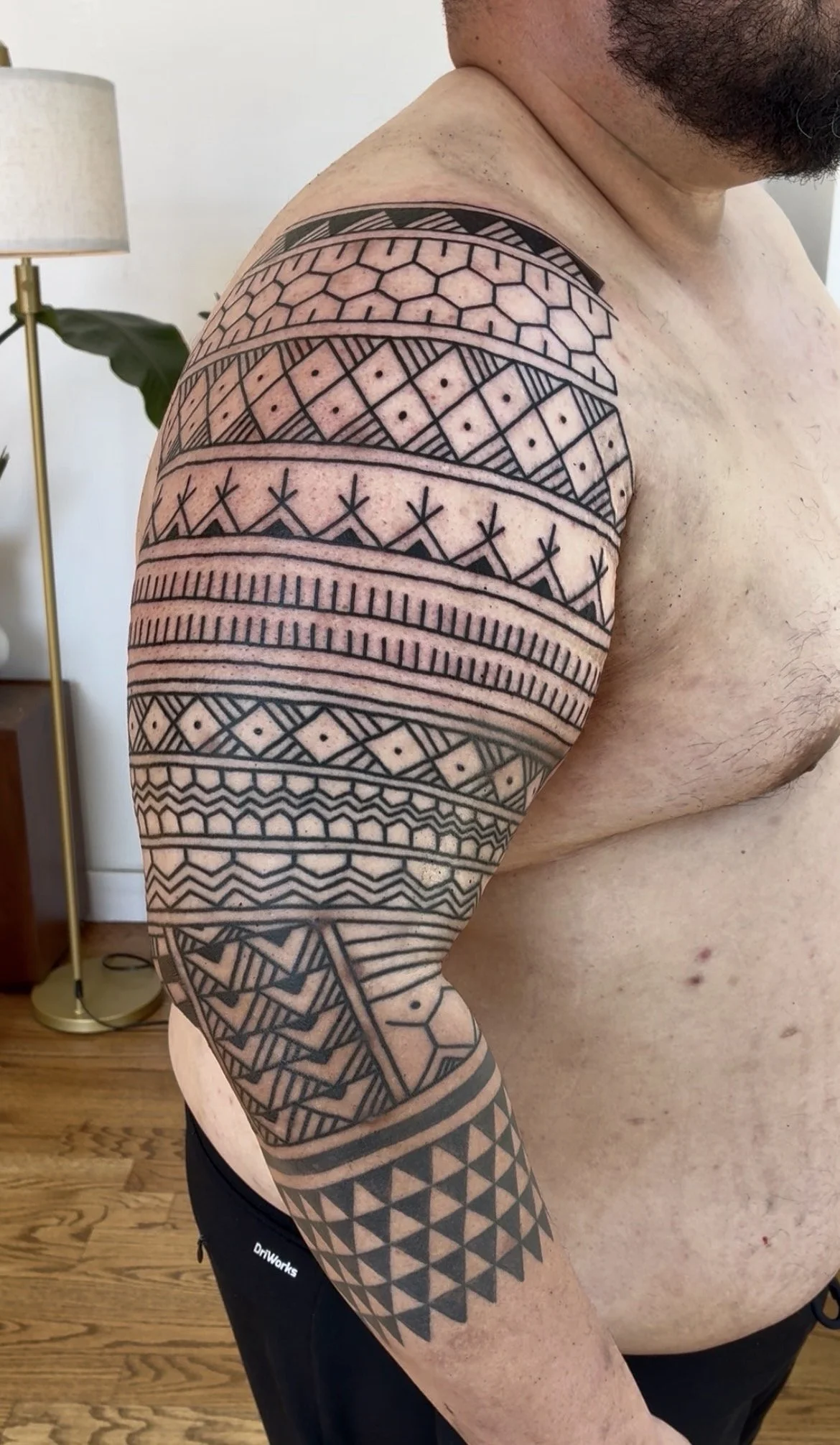 A person with a full sleeve tattoo featuring geometric patterns on their upper arm and shoulder, with a container of a plant and a standing lamp in a room background.