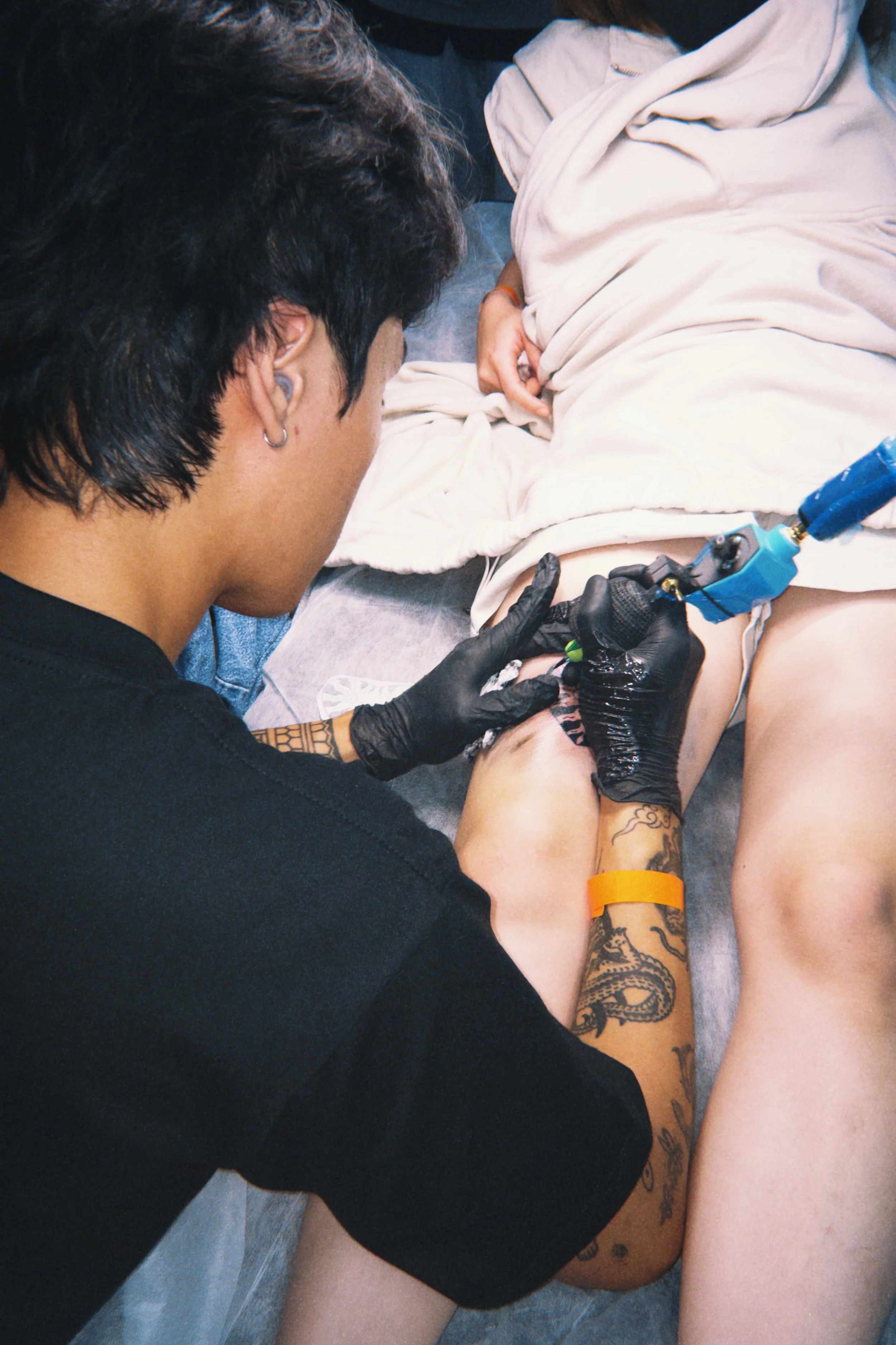 A person with tattoos on their arm, wearing black gloves, is tattooing another person lying down and wearing a white shirt and shorts.