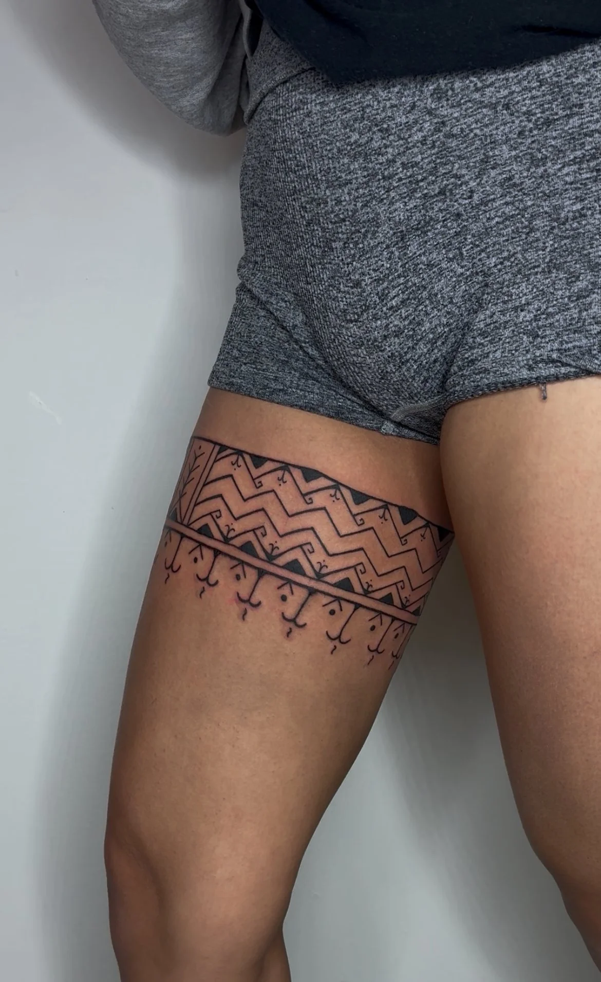 Close-up of a person's upper thigh and hips with a geometric black tattoo above the knee, wearing gray athletic shorts and a dark shirt, against a plain white wall background.