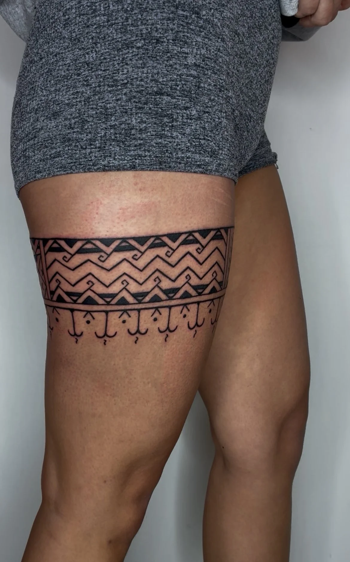 Close-up of a woman's upper thighs showing intricate black tattoo with geometric and tribal patterns, partially obscured by gray shorts.