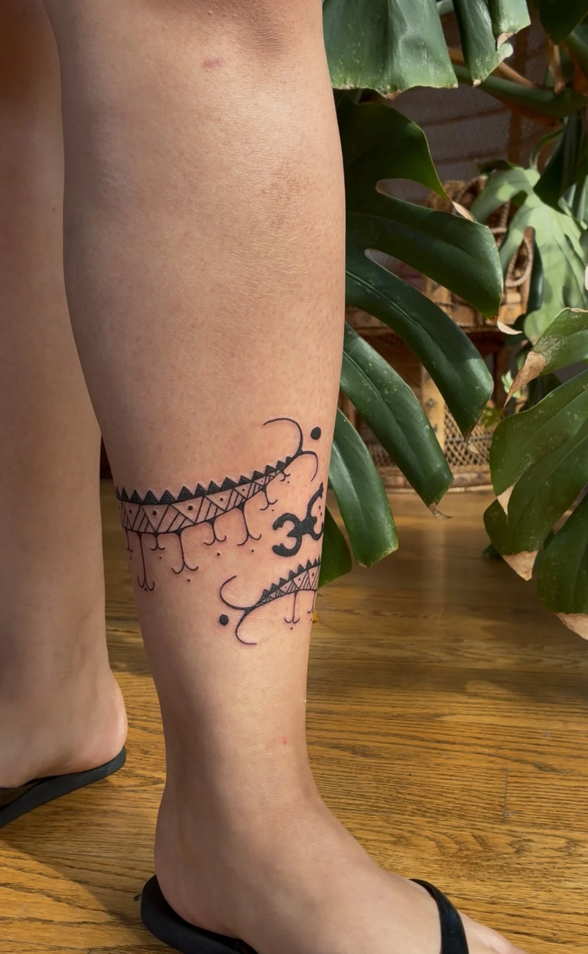A person's leg with a tattoo of intricate geometric and abstract designs, including the Om symbol, partially visible, next to green leafy plants on a wooden surface.