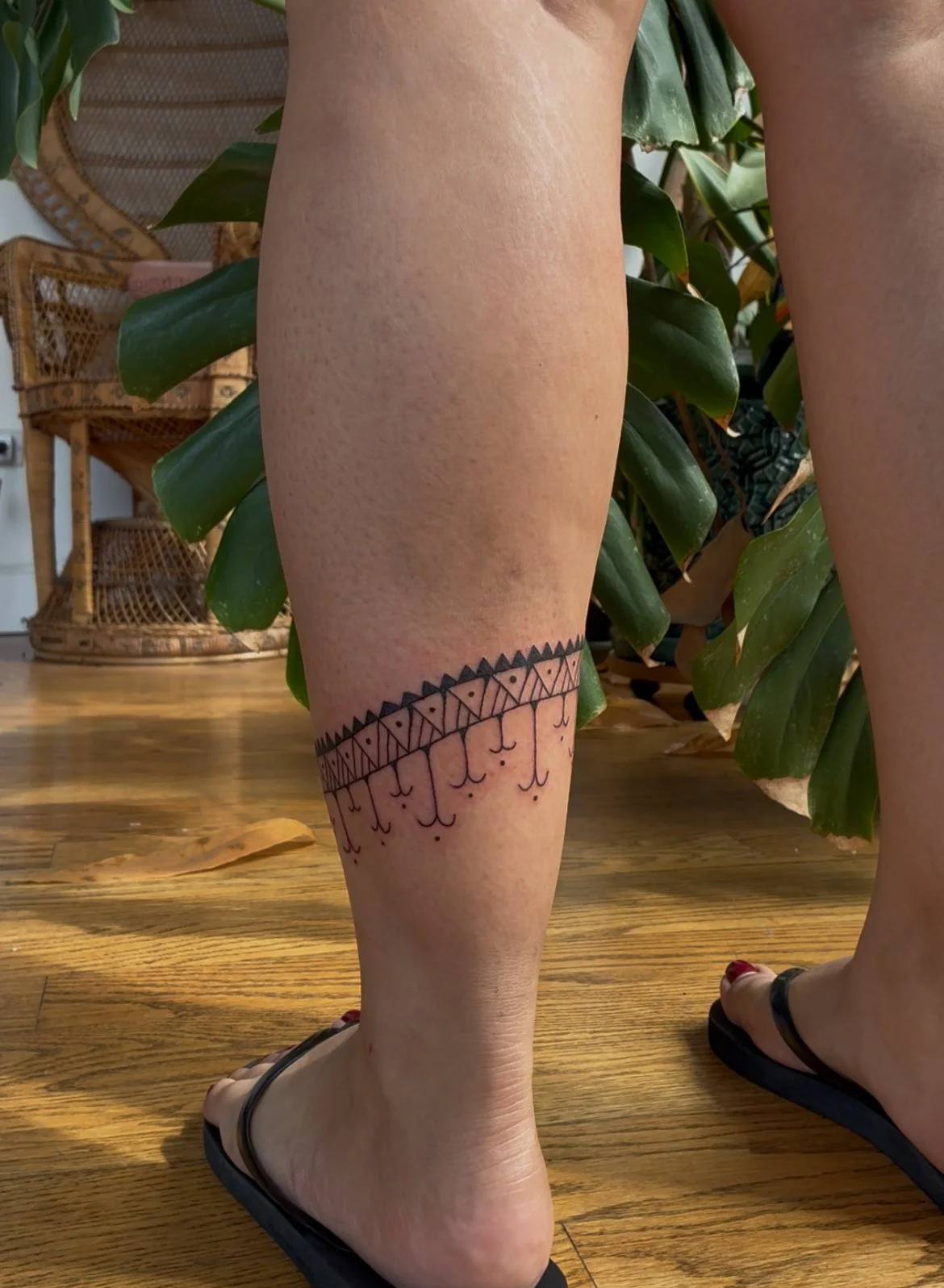 A person's lower leg with a black geometric tattoo design near the ankle, standing on a wooden floor with some green plants in the background.