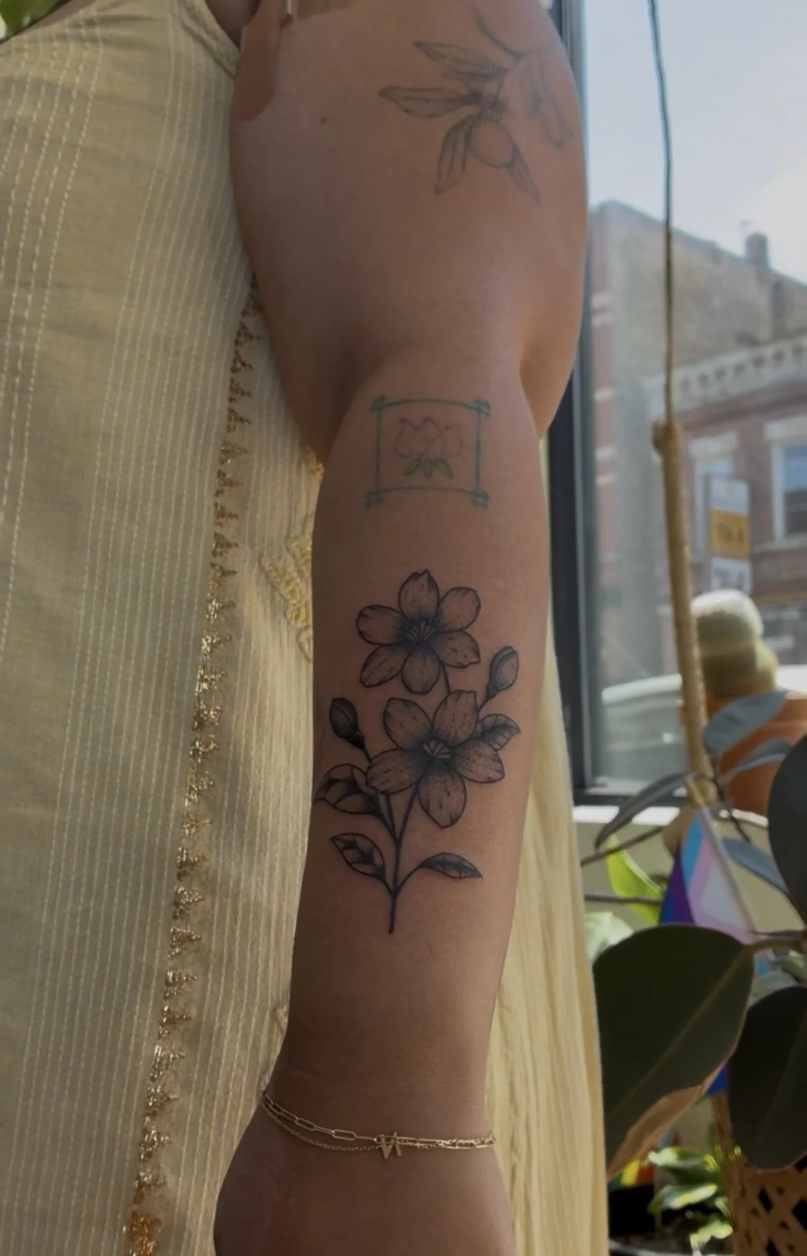 A person's arm with multiple tattoos including a flower, a framed flower, and a branch with leaves and fruit, with a gold bracelet and an anklet, standing indoors near a window with buildings outside.
