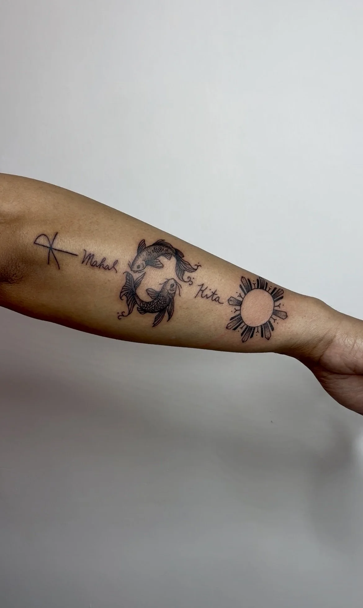 Tattoo of two koi fish swimming in a circle with the words 'Mahala' and 'Kikaa' written in cursive and a sun symbol with rays.