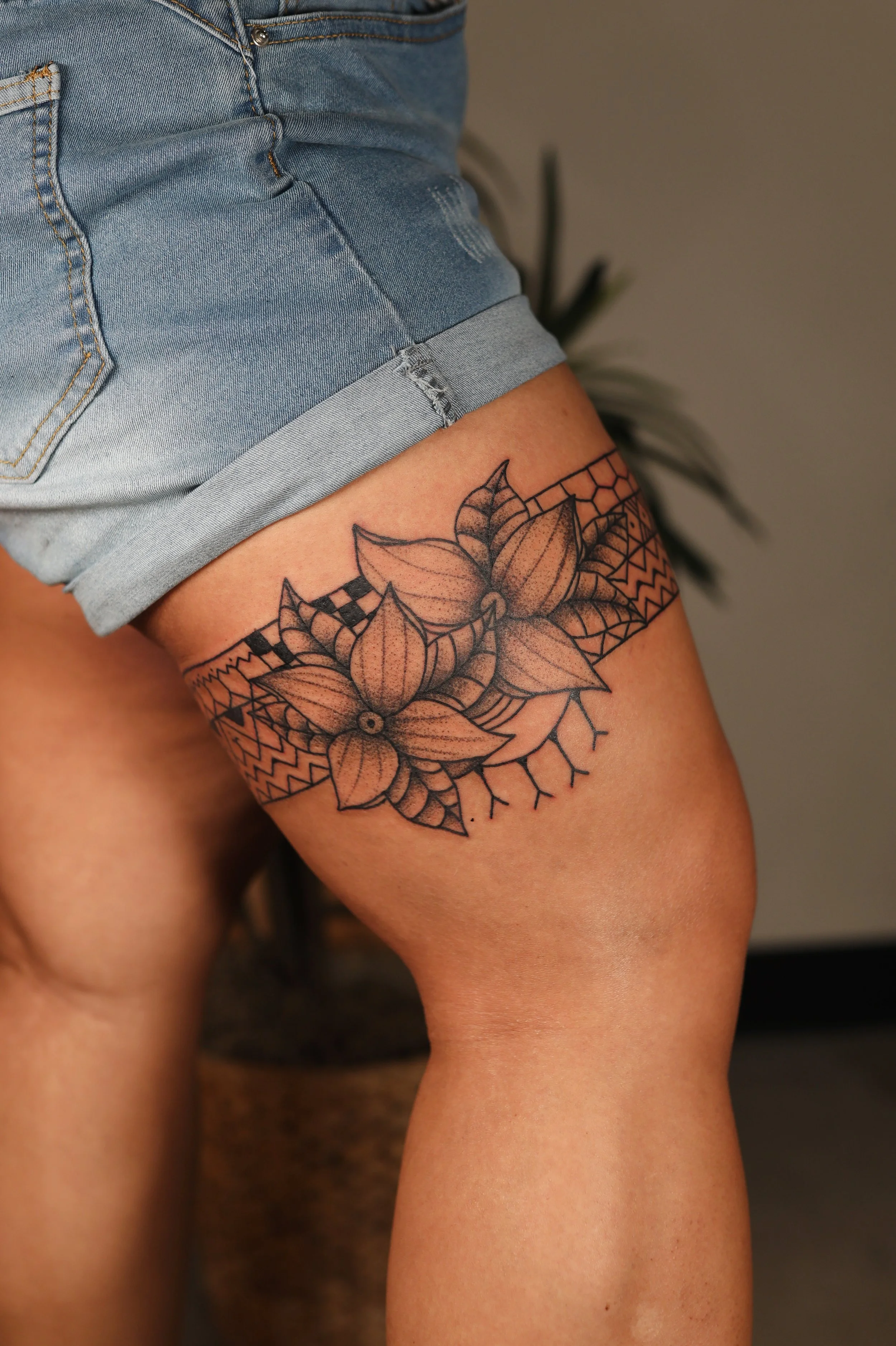 A close-up of a person's upper thigh showing a black ink floral tattoo with geometric details, wearing light denim shorts with a rolled hem.