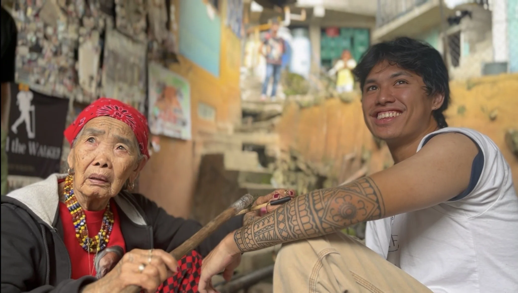 A young man with tattoos and a white tank top smiling and holding hands with an elderly woman dressed in traditional clothing and jewelry, sitting together outdoors in a colorful, rustic village setting.