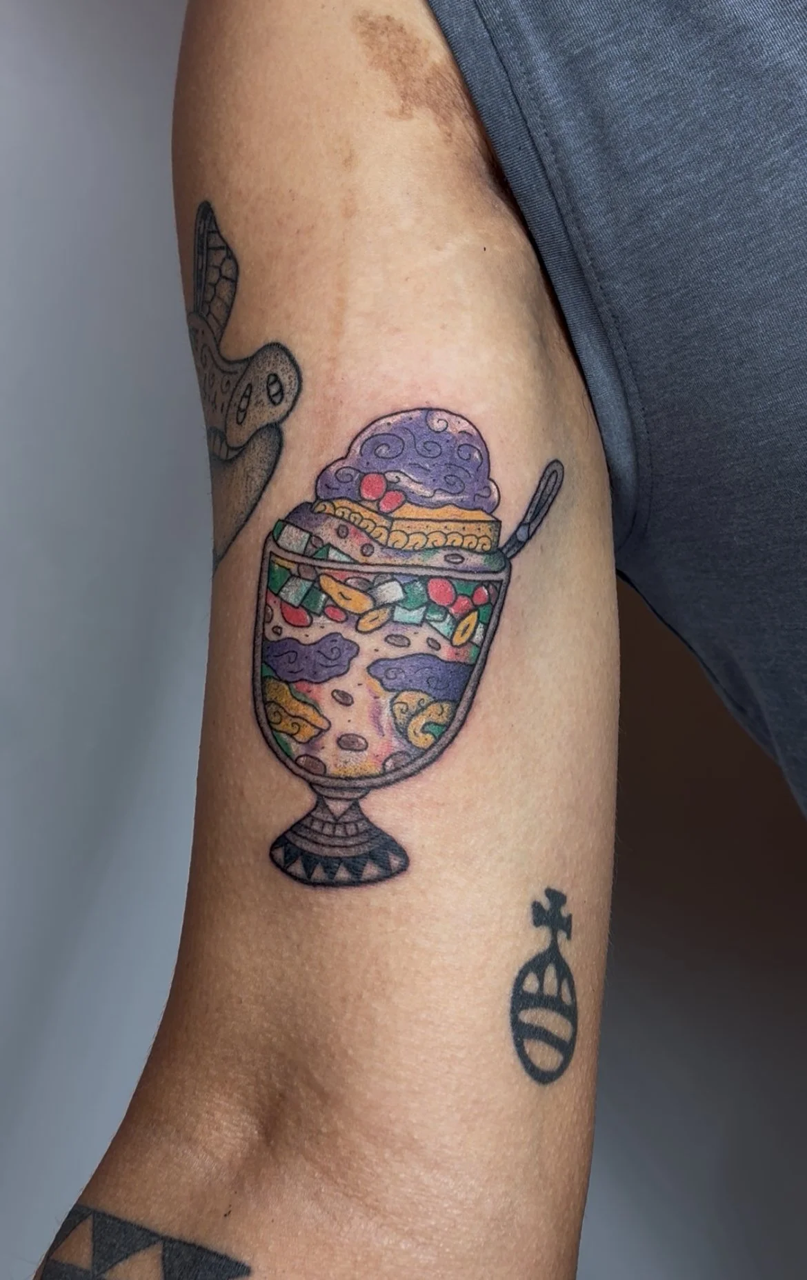 Tattoo of a colorful halo halo desert mountain glass with a purple sundae on top, featuring various swirls and toppings, on a person's upper arm.