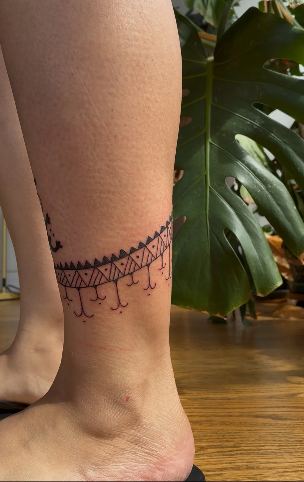 Close-up of a person's ankle with a black geometric tattoo around the ankle and small line designs hanging down, with a large green leafy plant in the background.
