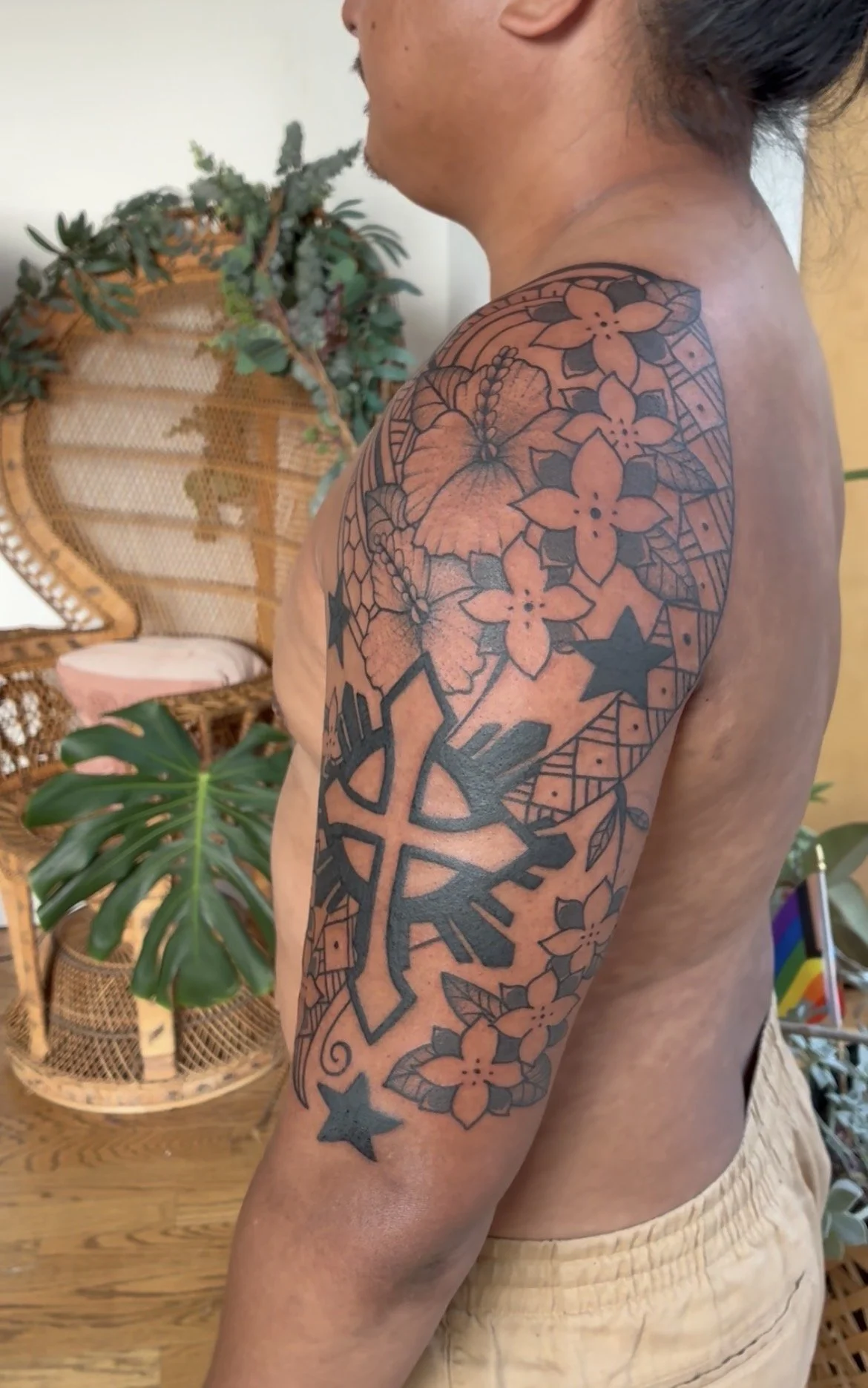 Side view of a shirtless person with a large floral tattoo on their upper arm and shoulder, standing indoors near a wicker chair with plants in the background.