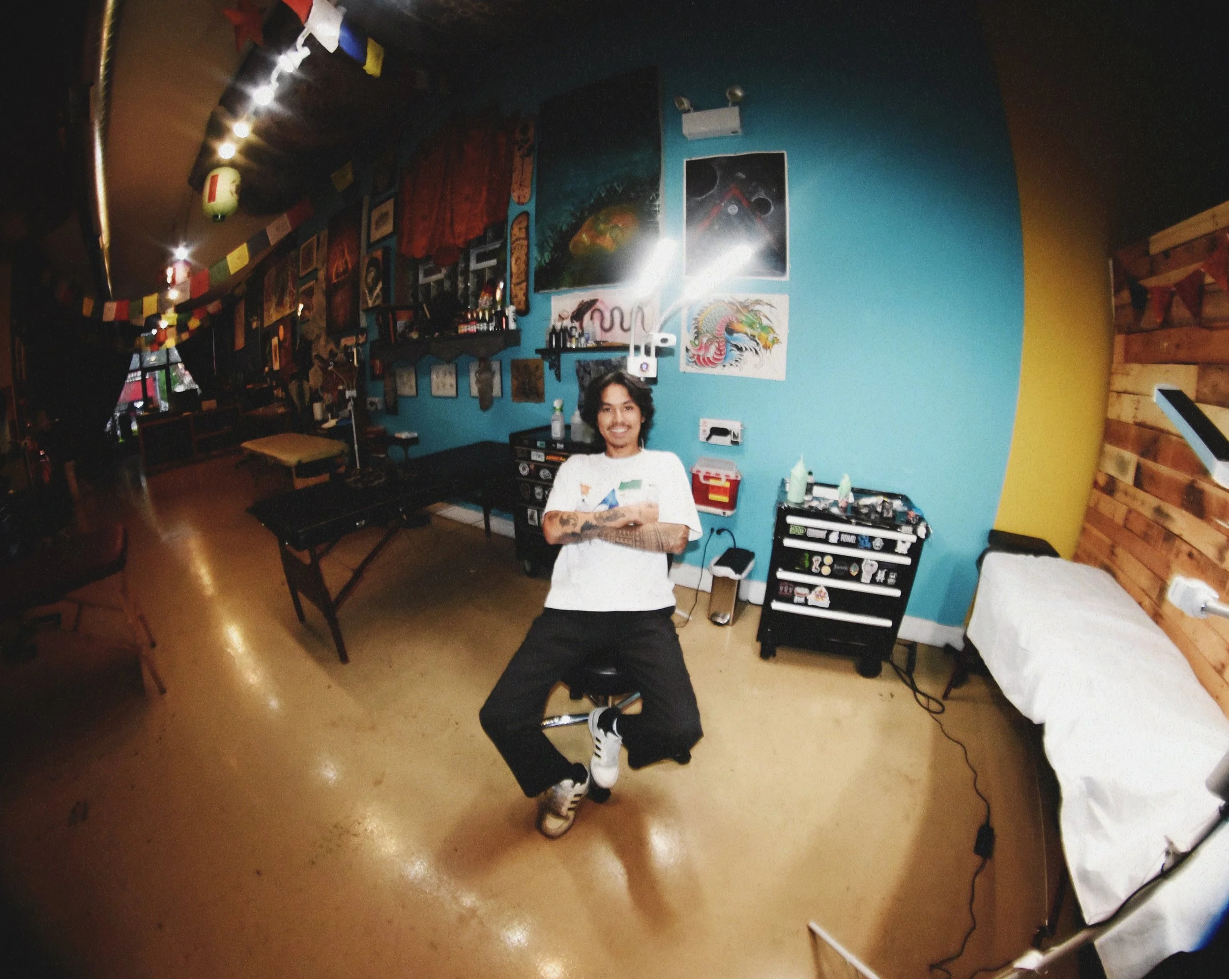 A young man with tattoos on his arms and leg, wearing a white T-shirt and black pants, standing in a room with colorful graffiti art and decorations, smiling at the camera.