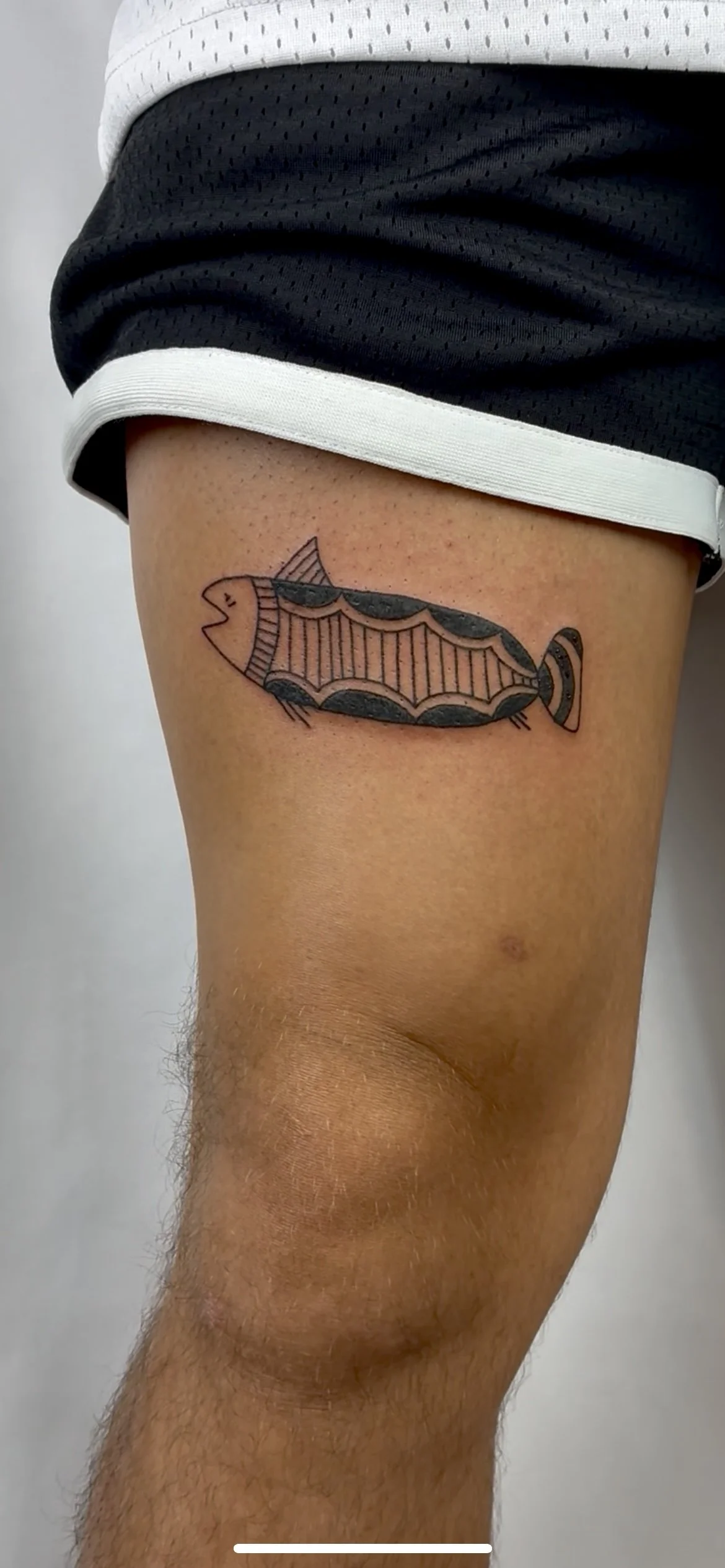 Tattoo of a fish with a geometric pattern on the thigh, wearing a smile, with a black, yellow, and orange color scheme.