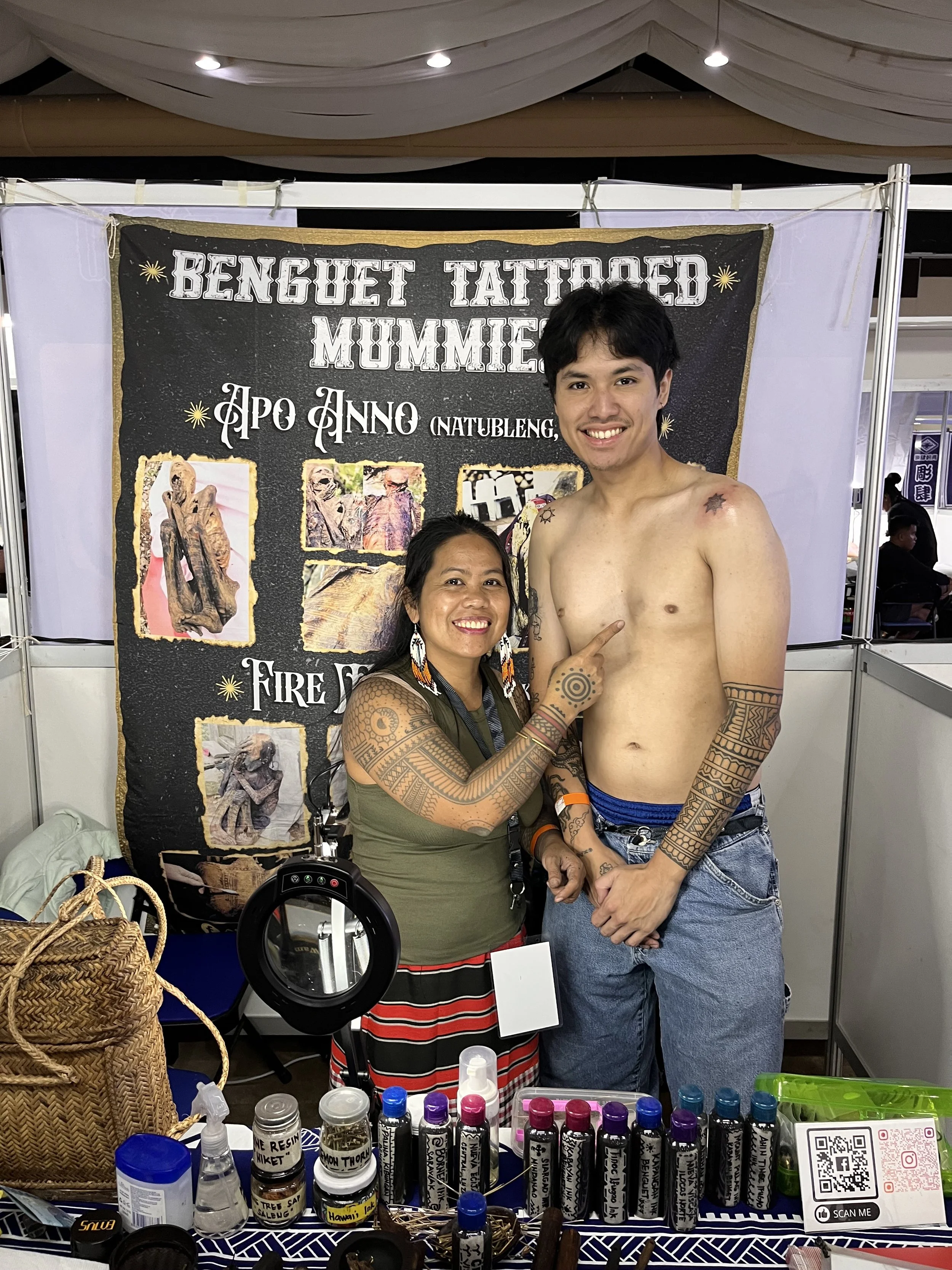 A woman and a shirtless man with tattoos standing in front of a display booth for Benguet Tatted Mummies at a fair or exhibition. The booth has various bottles of paint or ink, a basket, and other craft supplies, with a black banner behind featuring images and text.
