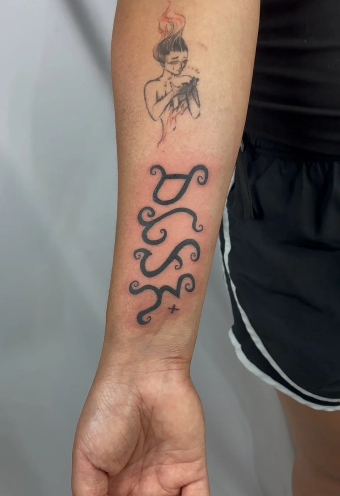 Tattoo on person's forearm depicting a girl with short hair, smoking a cigarette, with fire or smoke coming from her head. Below the girl, the word 'YES' is written in large, decorative script, followed by a small cross.