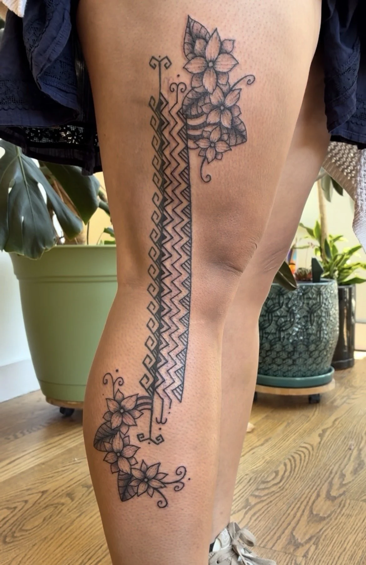 A tattoo on a person's thigh features a geometric pattern interwoven with floral designs, including flowers and leaves, in black ink.