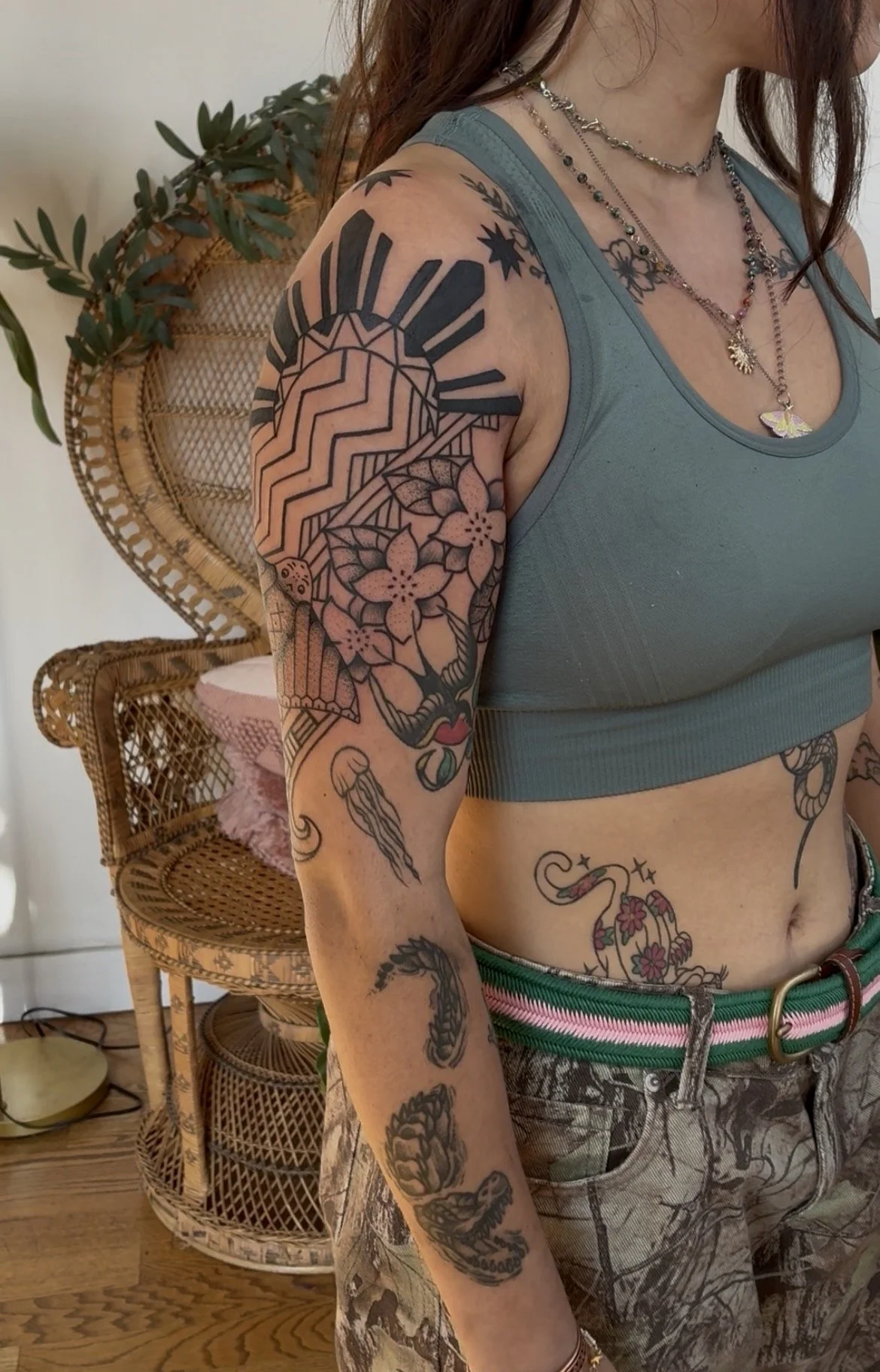 A woman with multiple tattoos on her arm and torso, wearing a teal sports crop top, layered necklaces, and patterned shorts, standing in a room with wooden furniture and a plant in the background.