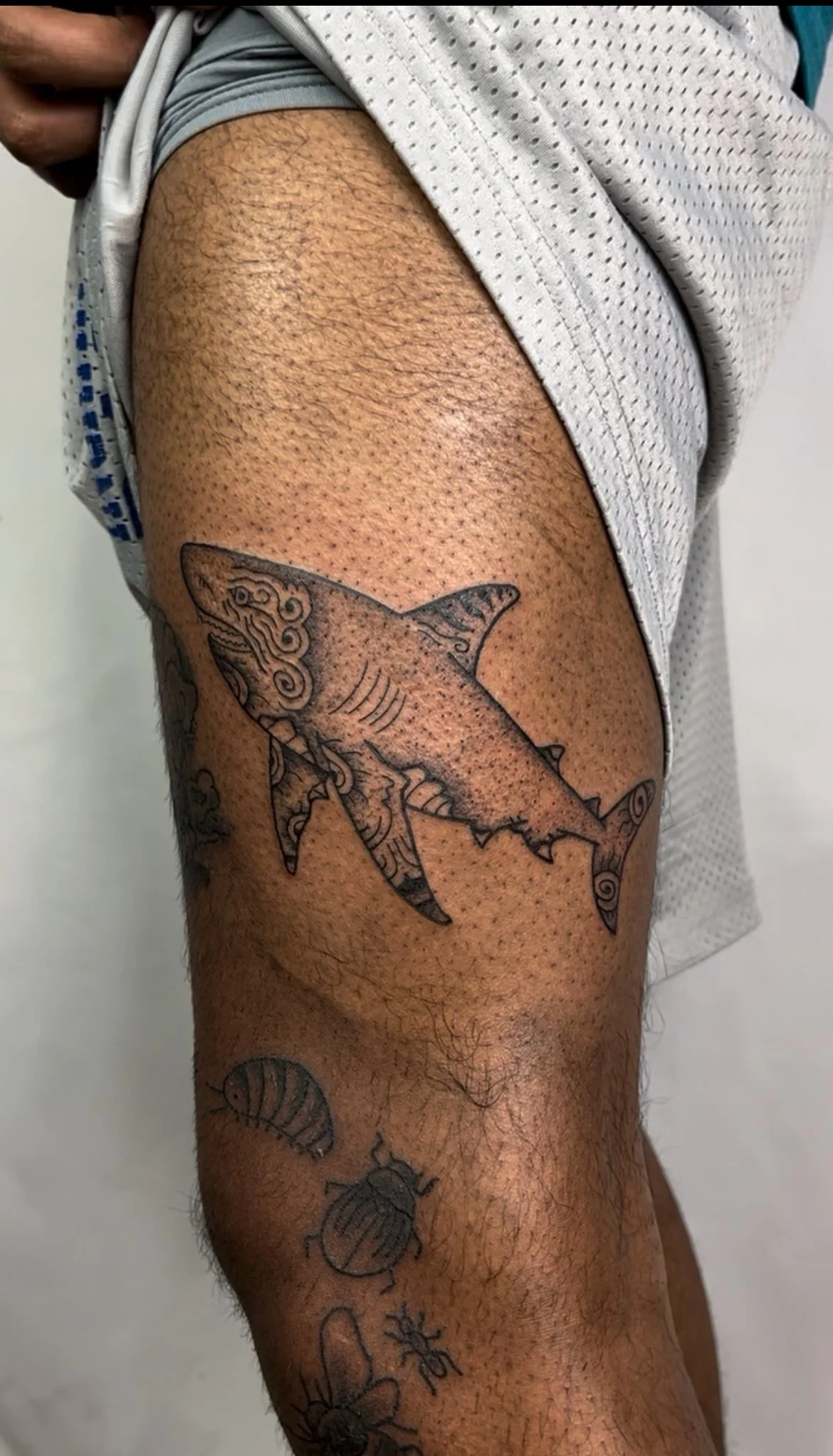 Tattoo of a stylized shark on a person's thigh, with other smaller tattoos including a bee, a ladybug, a flower, and other small insects.