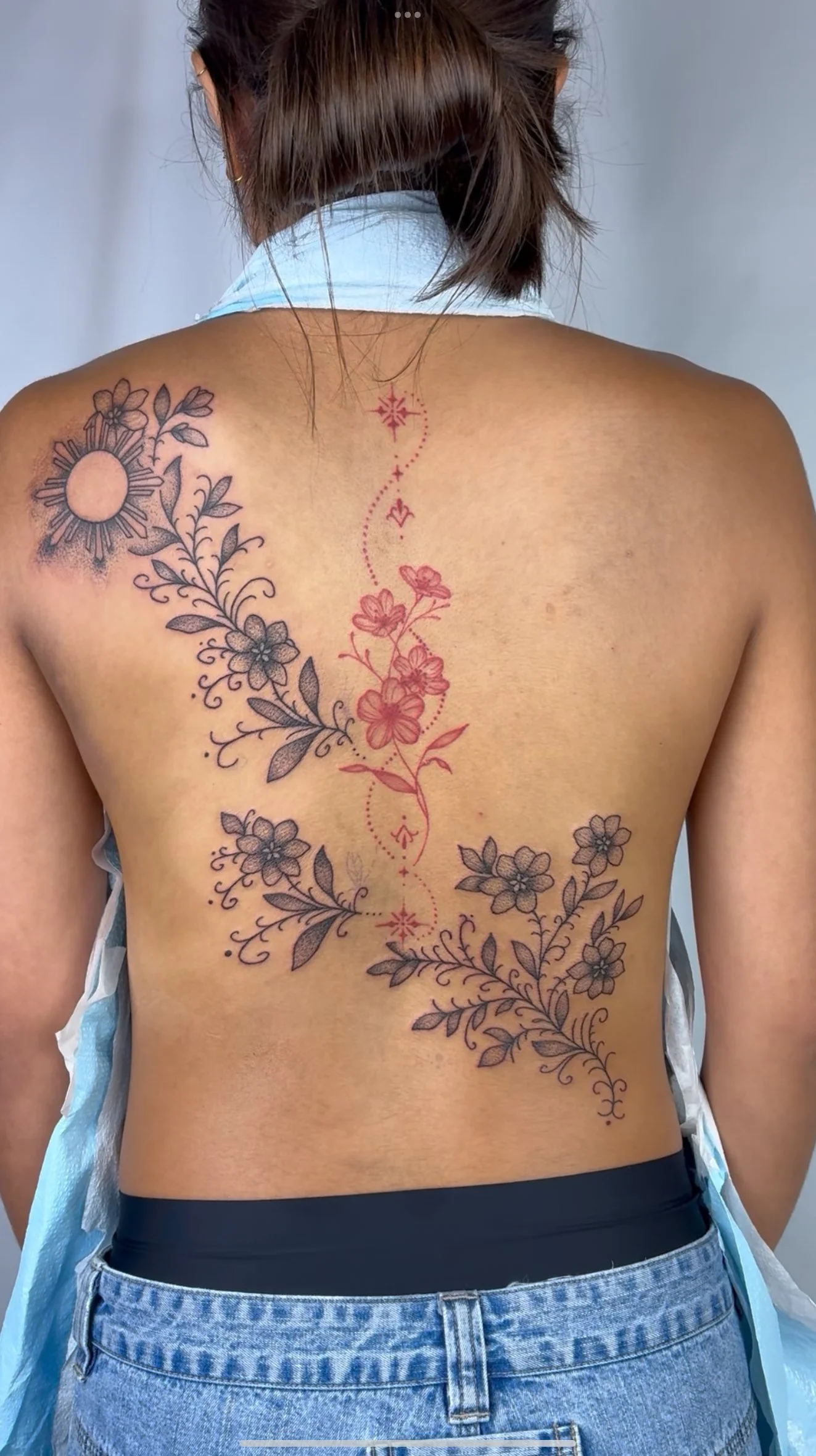 Back view of a person with a floral tattoo on their back, featuring flowers and leaves in black and gray, with a decorative red vertical line running down the center.