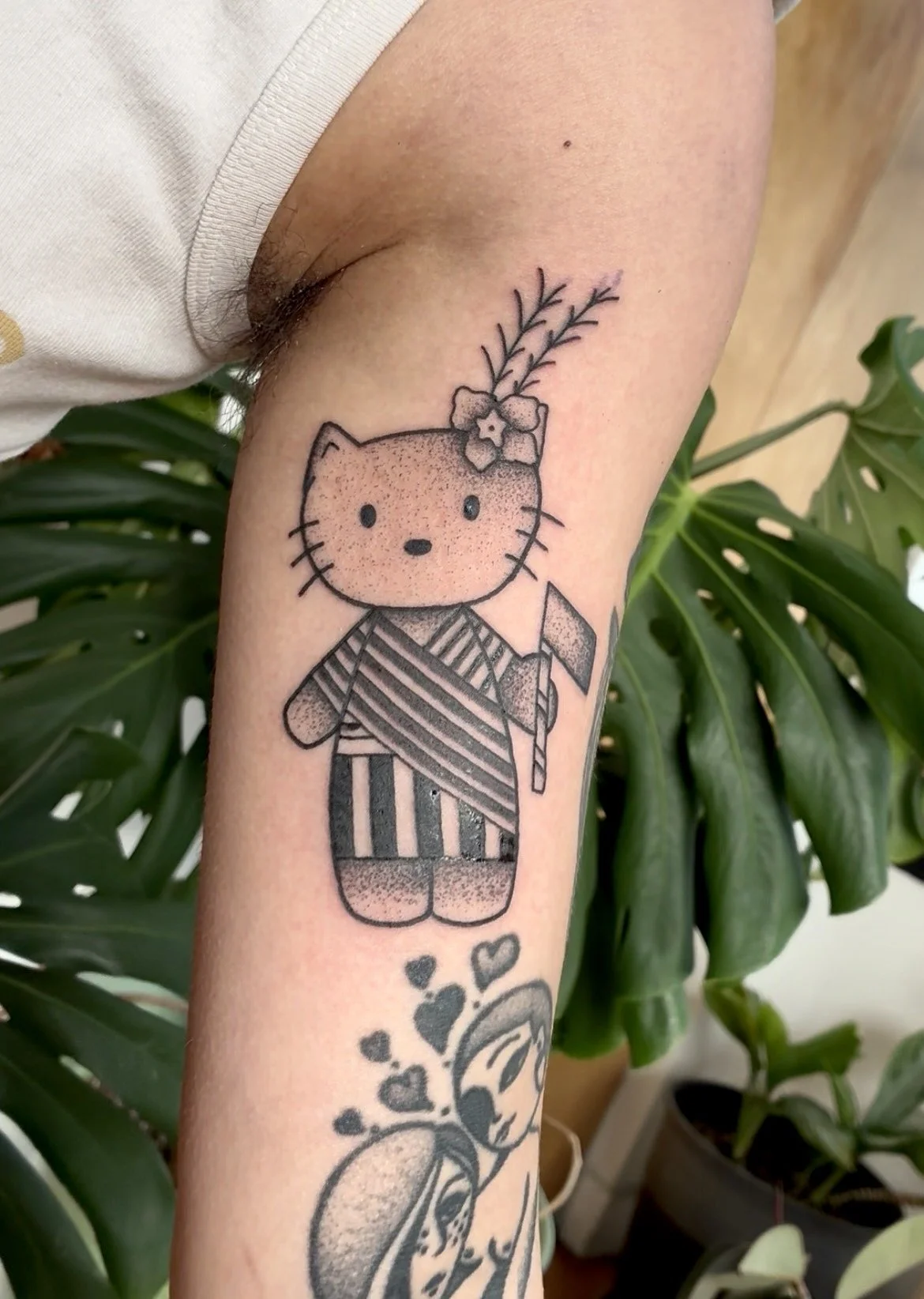 Tattoo of a cartoon cat dressed in traditional Japanese kimono, holding a flag, with a small flower and plant above its head, on a person's forearm.