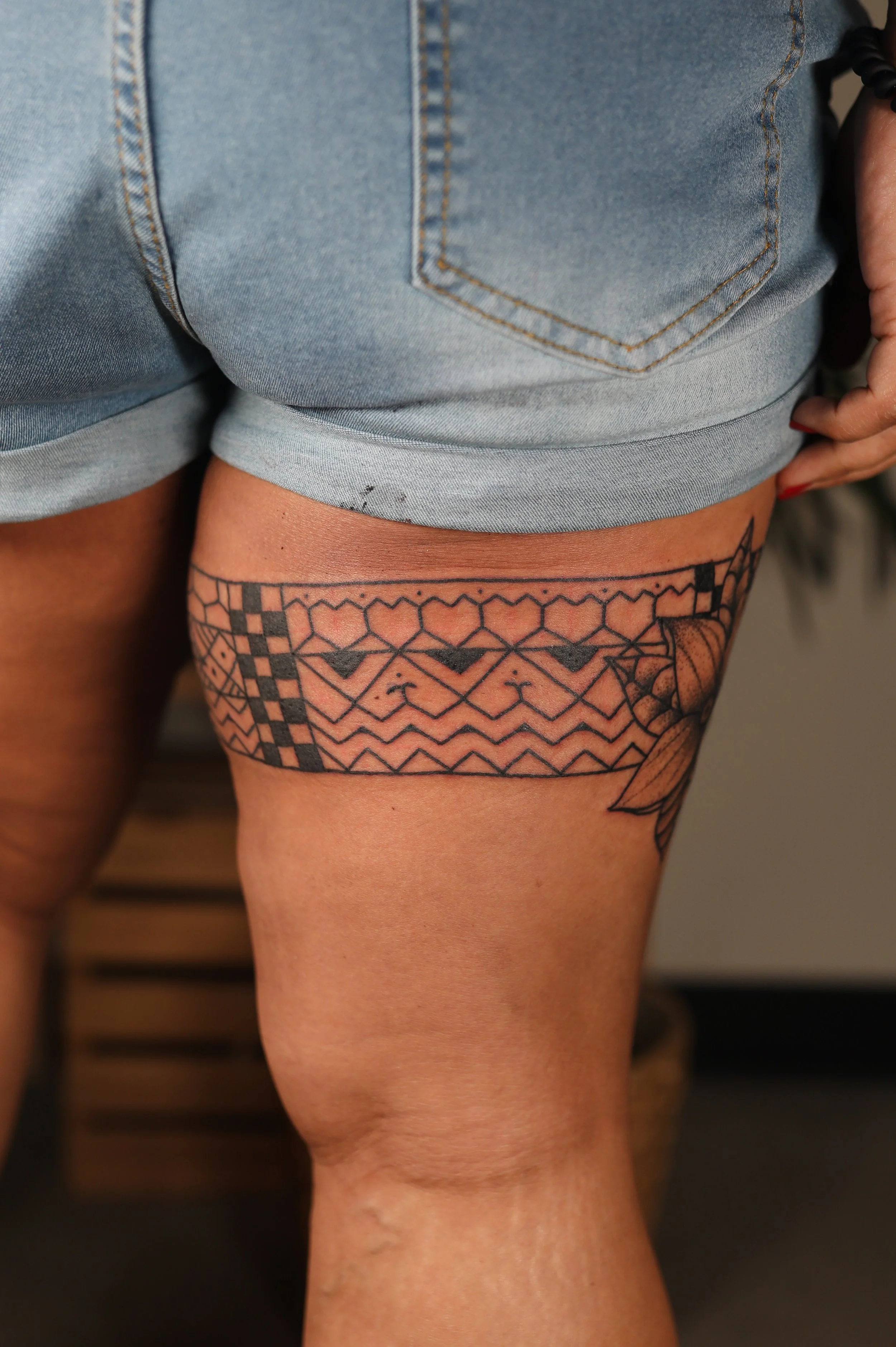 Close-up of a person's upper thigh showing a tattoo with geometric patterns, surrounded by denim shorts.