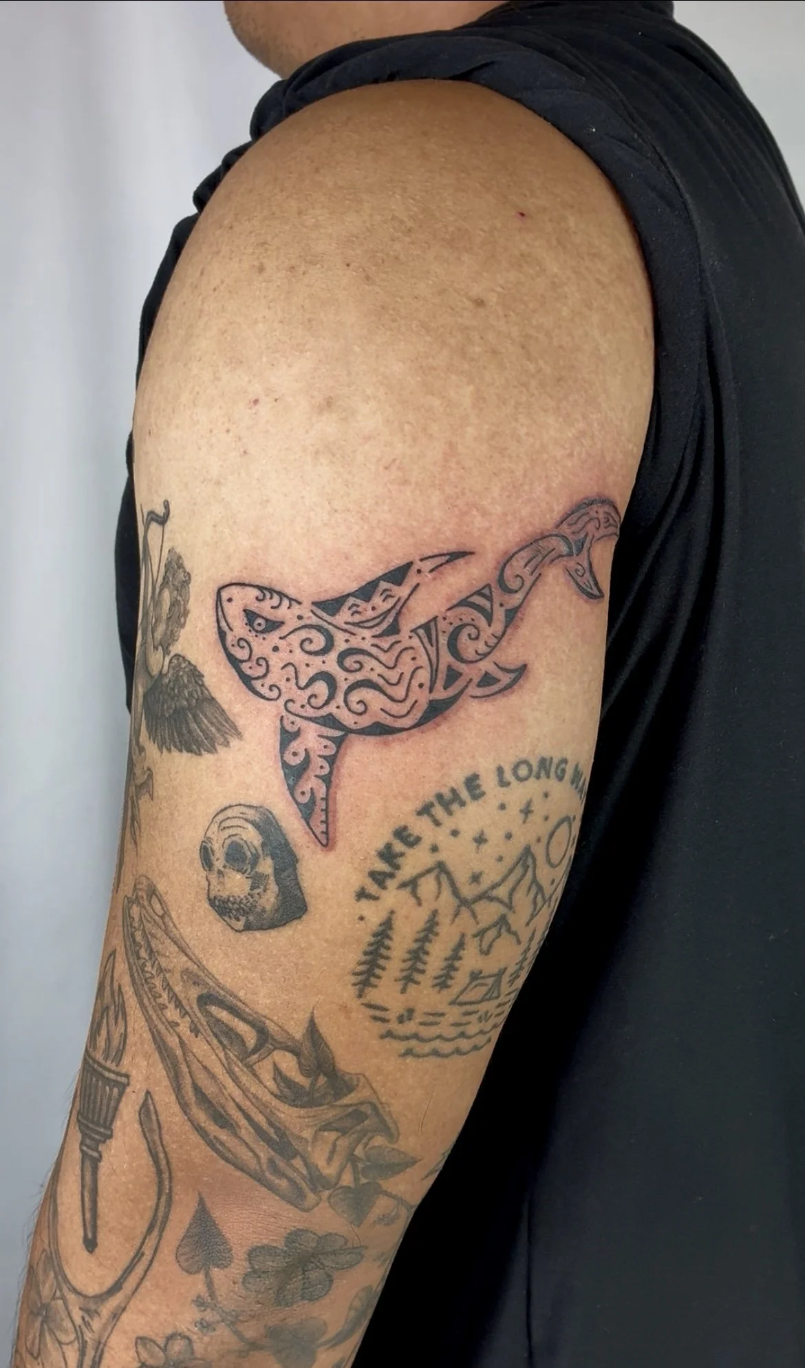 Close-up of a tattooed person's upper arm, featuring a stylized whale with intricate patterns, a skull, a feather, and other designs.