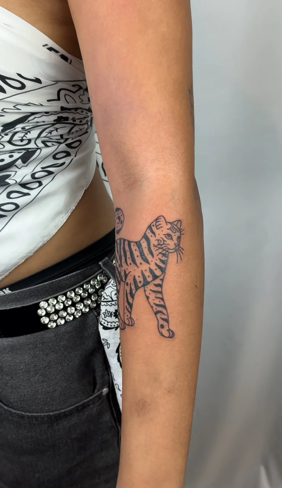 Close-up of a person's arm with a tattoo of a tiger, wearing black jeans with a rhinestone belt, and a white bandana with black patterns.