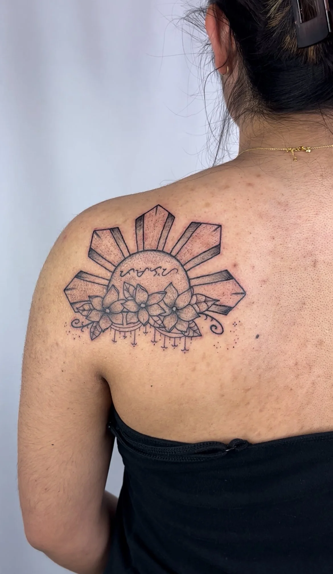 Tattoo on woman's shoulder featuring a geometric sun with rays, a circular element with scribbled text, and a floral design with lilies and hanging ornaments.