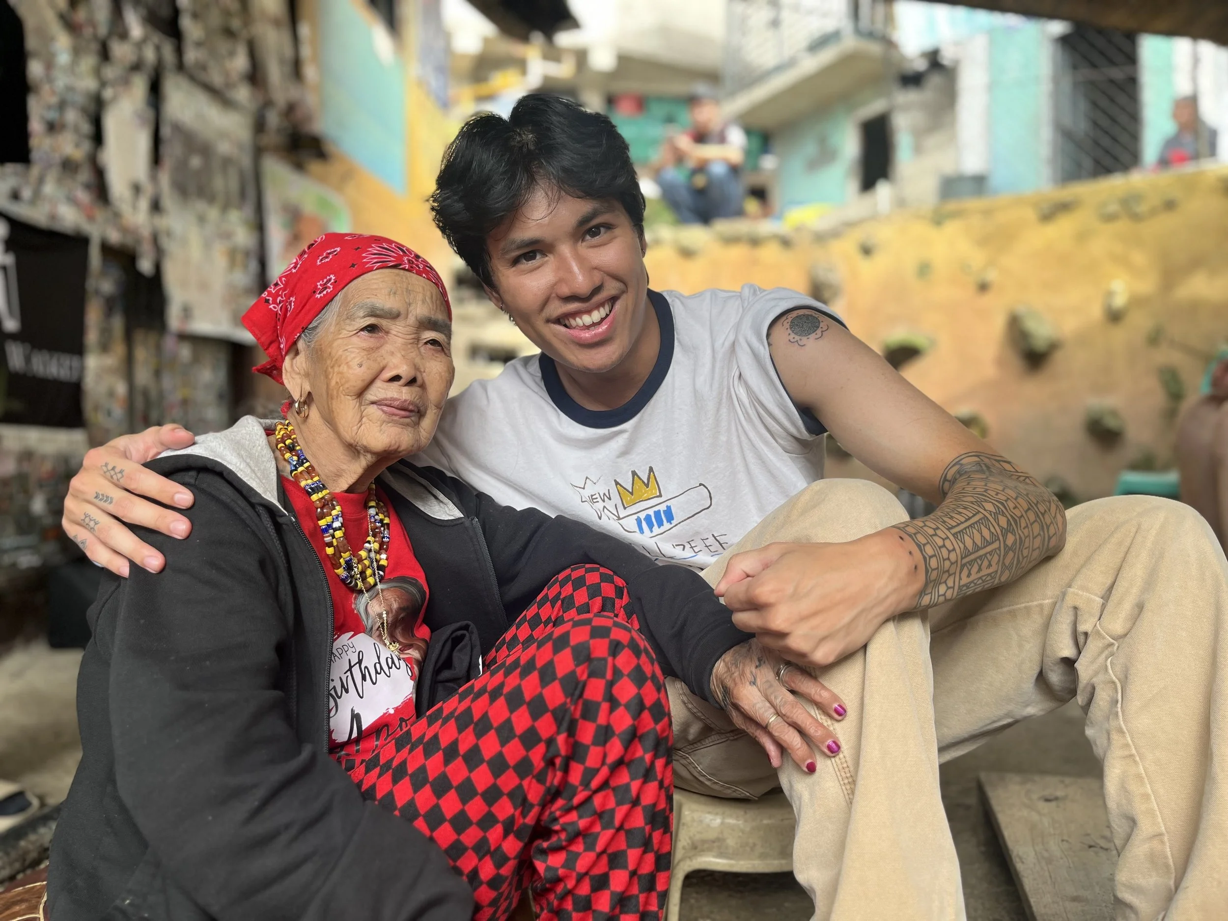 A young man with dark hair and tattoos on his arms taking a selfie with an elderly woman with gray hair, wearing a red bandana and colorful jewelry, both smiling. They are outdoors in a vibrant, urban environment with colorful walls and murals in the background.