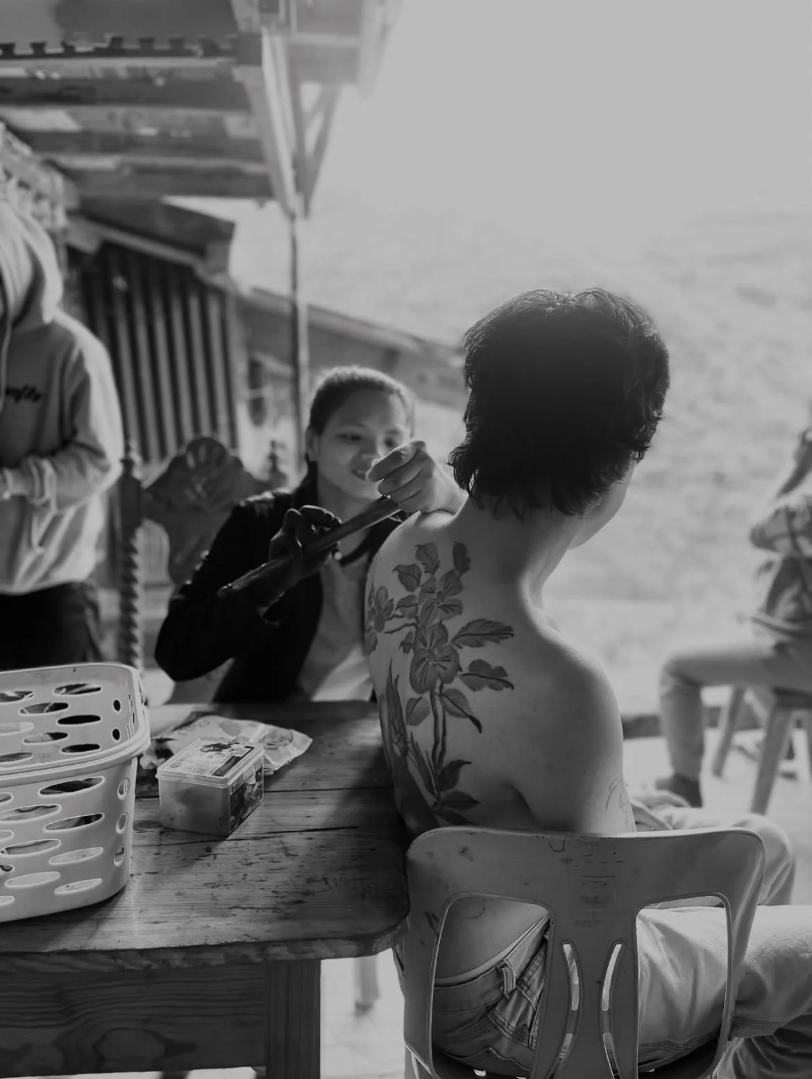 A person with a floral tattoo on their back sitting at a table while a woman with a smile paints a large floral design on their shoulder, in a casual outdoor setting.