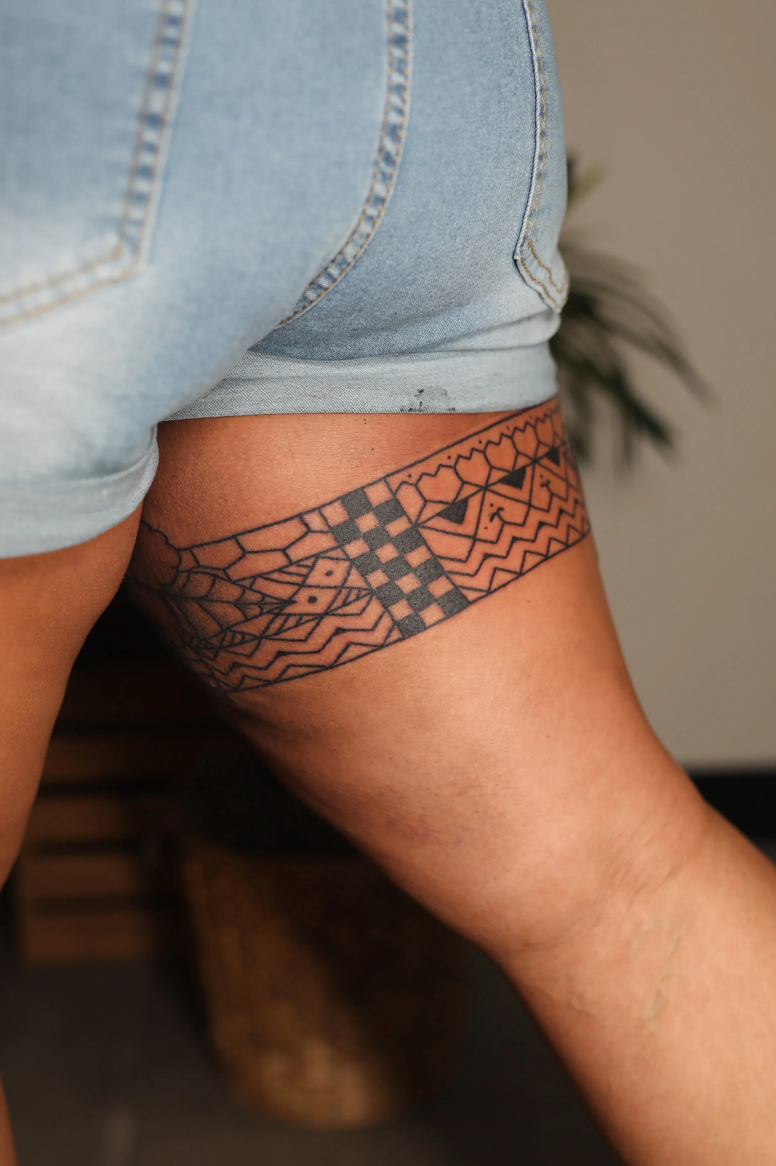 Close-up photo of a person's upper thigh and hip, showing a black tribal tattoo on the skin beneath a light blue denim short with visible stitching.
