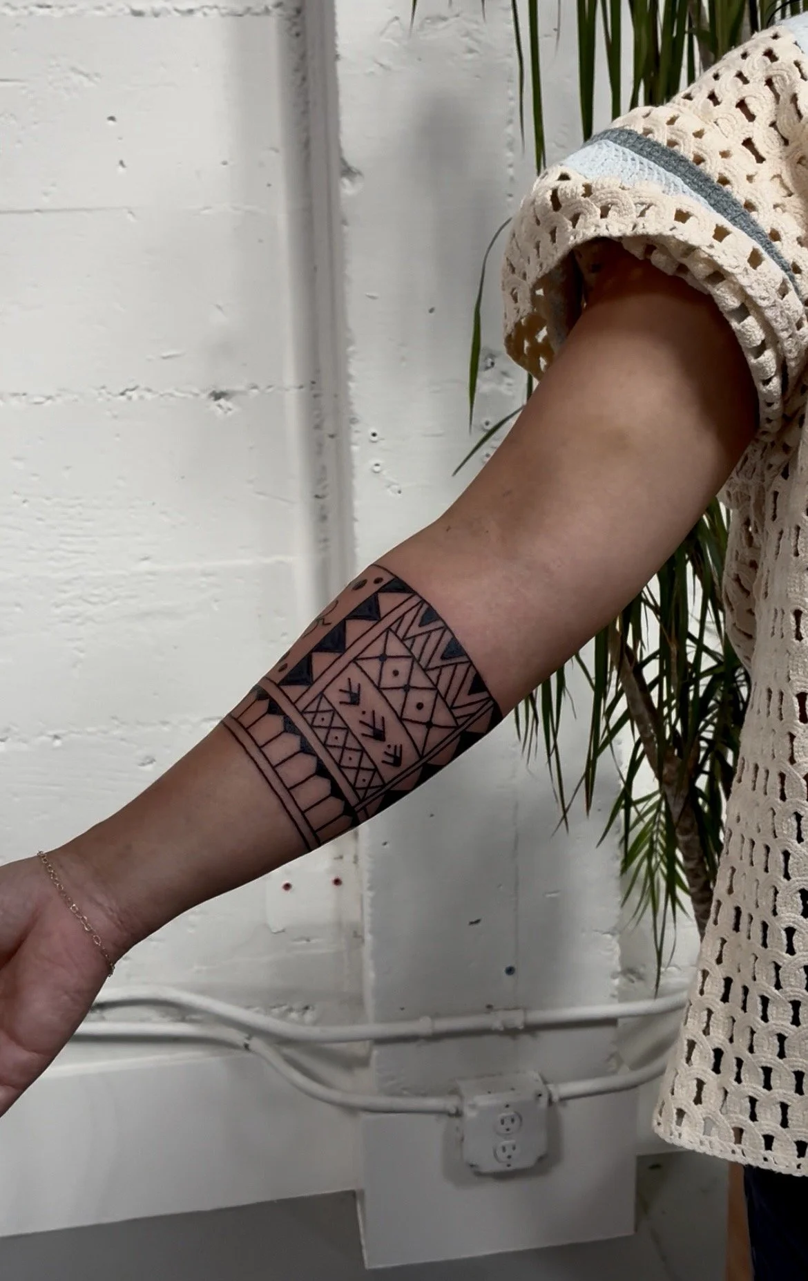 A person’s arm with a geometric tribal-style tattoo, wearing a beige crochet short sleeve top, standing in front of a white brick wall and some green plants.