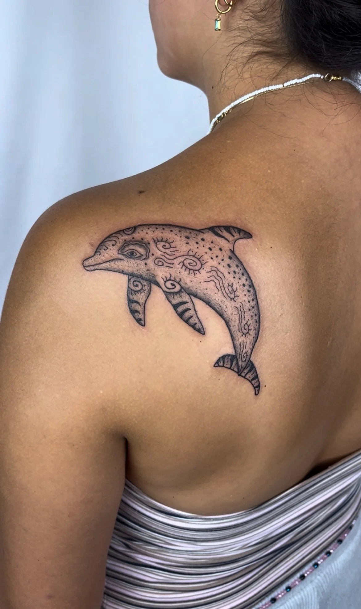 A person's shoulder features a black ink tattoo of a dolphin with decorative swirls and dots, located on the upper arm near the shoulder.