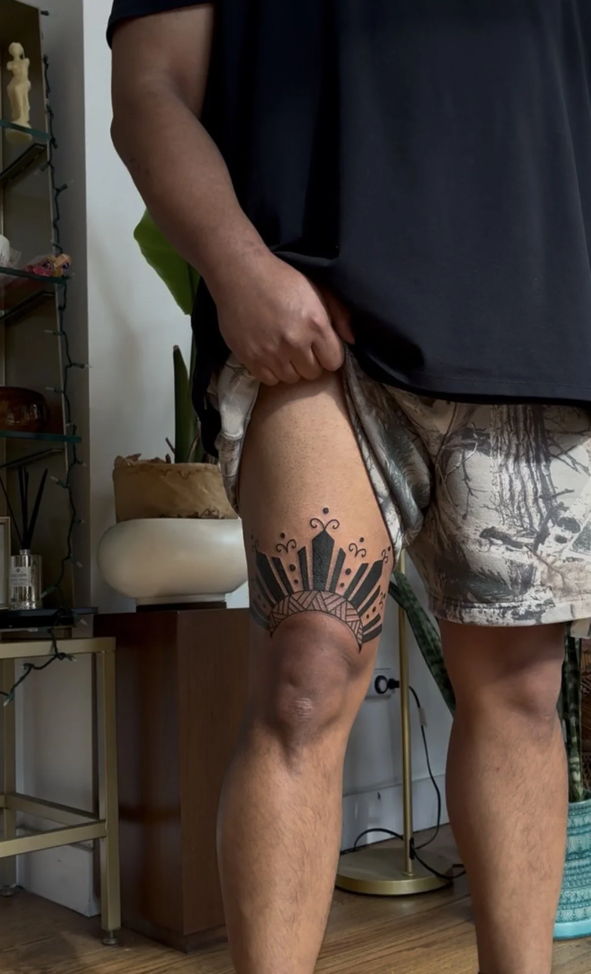 A man wearing black t-shirt and camo shorts with a tattoo on his thigh, standing indoors with a plant, a white chair, and a shelving unit in the background.
