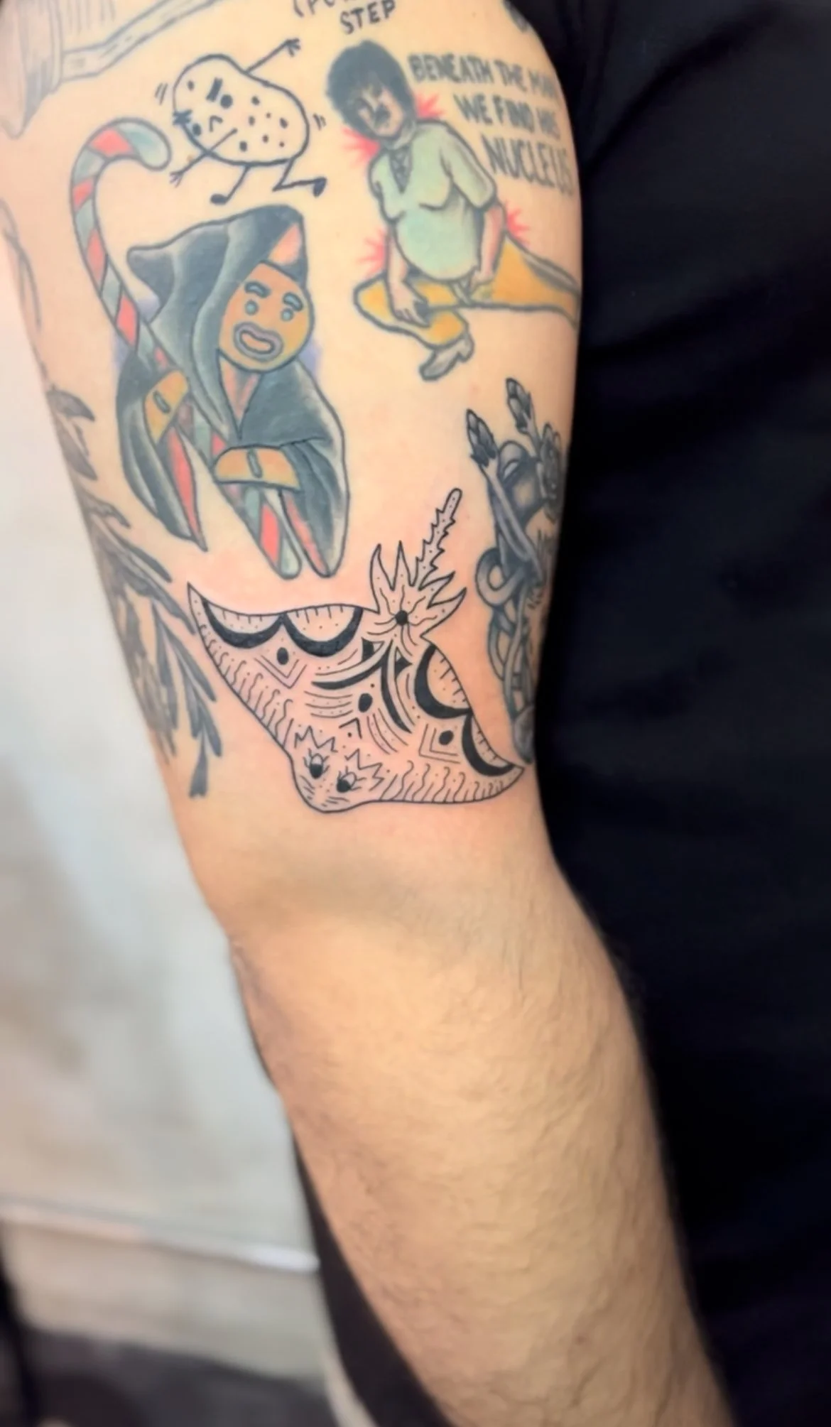 Tattoo on person's arm depicting various cartoon-style characters and designs, including a potato, a hooded figure, and a sleeping animal with intricate patterns.