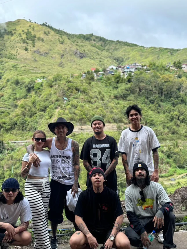 Group of six people outdoors on a hillside with green vegetation and houses in the background.