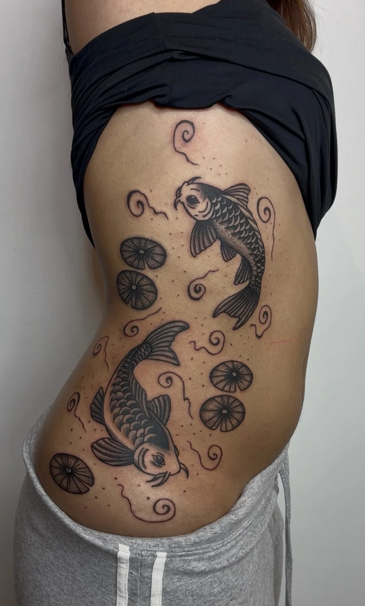 Tattoo of two koi fish swimming among lily pads and swirling water patterns on person's side.