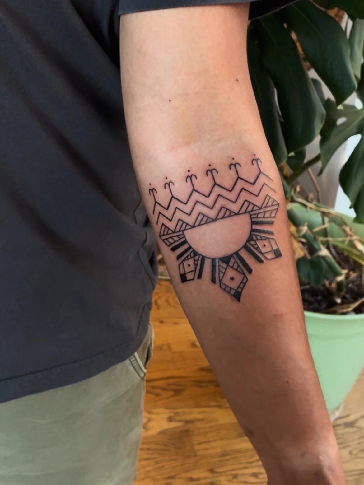 A geometric black tattoo on a person's forearm, featuring a semi-circle with rays extending outward and a patterned border around it, with small symbols above.