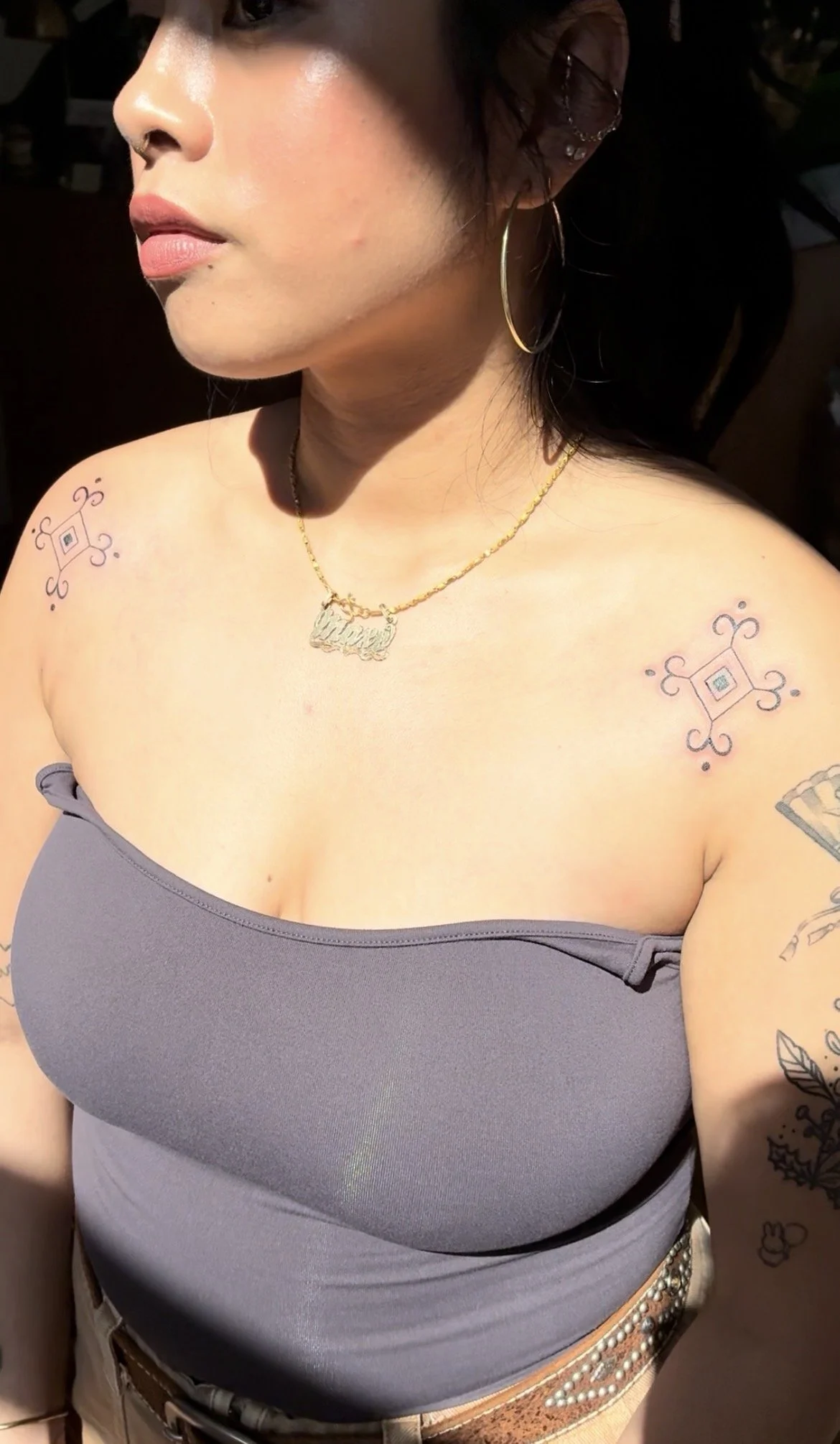Close-up of a woman with tattoos on shoulders and arm, wearing hoop earrings, a necklace with a nameplate, a gray strapless top, and beige pants with a bejeweled belt.