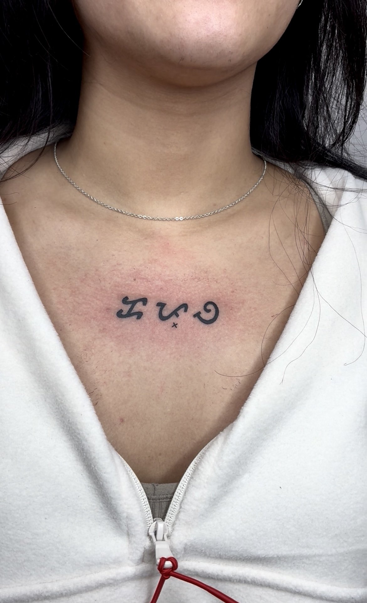 Close-up of a person's neck and upper chest showing a thin silver necklace and a tattoo with stylized  baybayin characters.