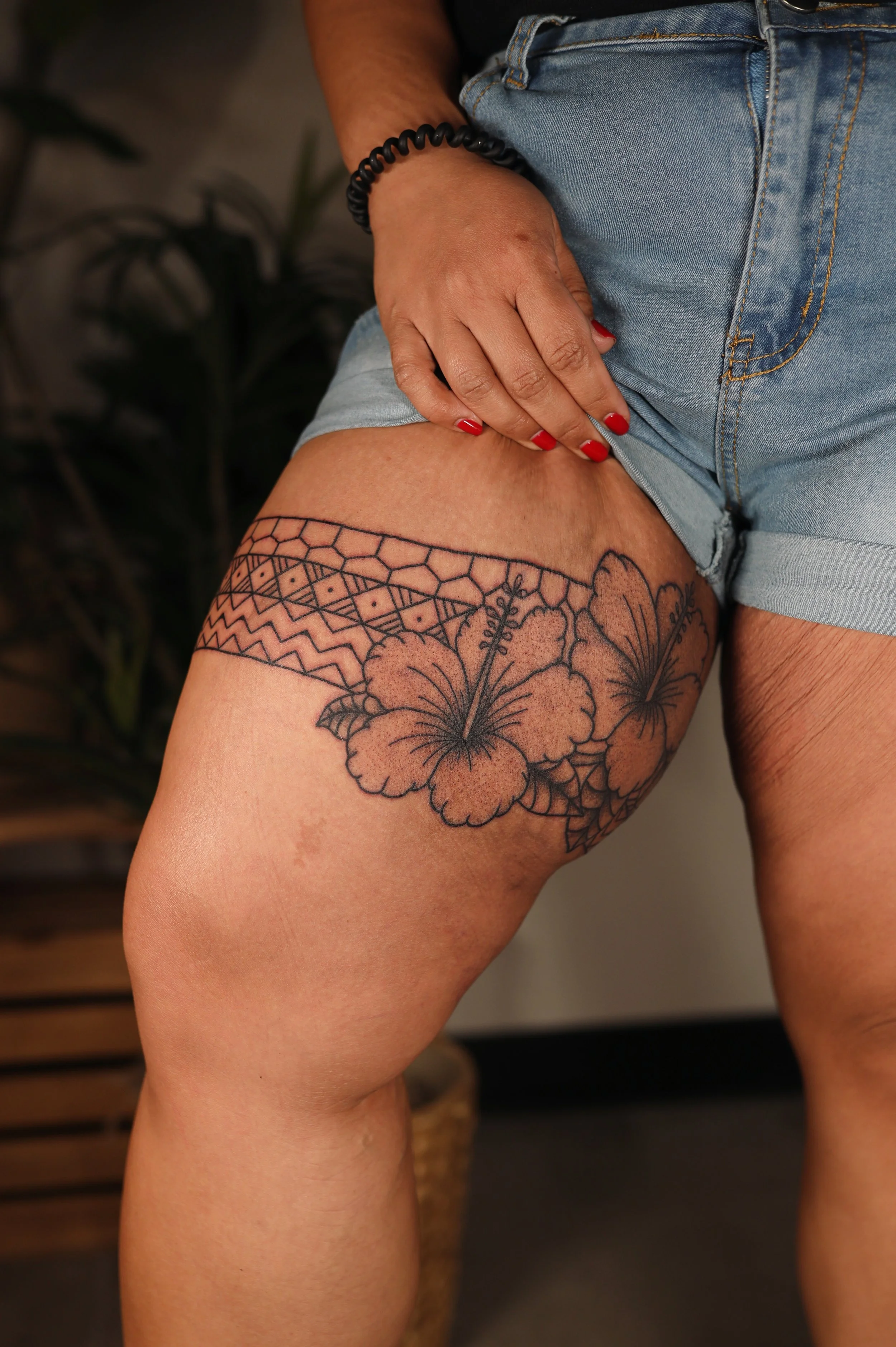 A woman wearing denim shorts with a floral tattoo on her upper thigh, red nail polish, and a black hair tie on her wrist, standing indoors with blurred background.