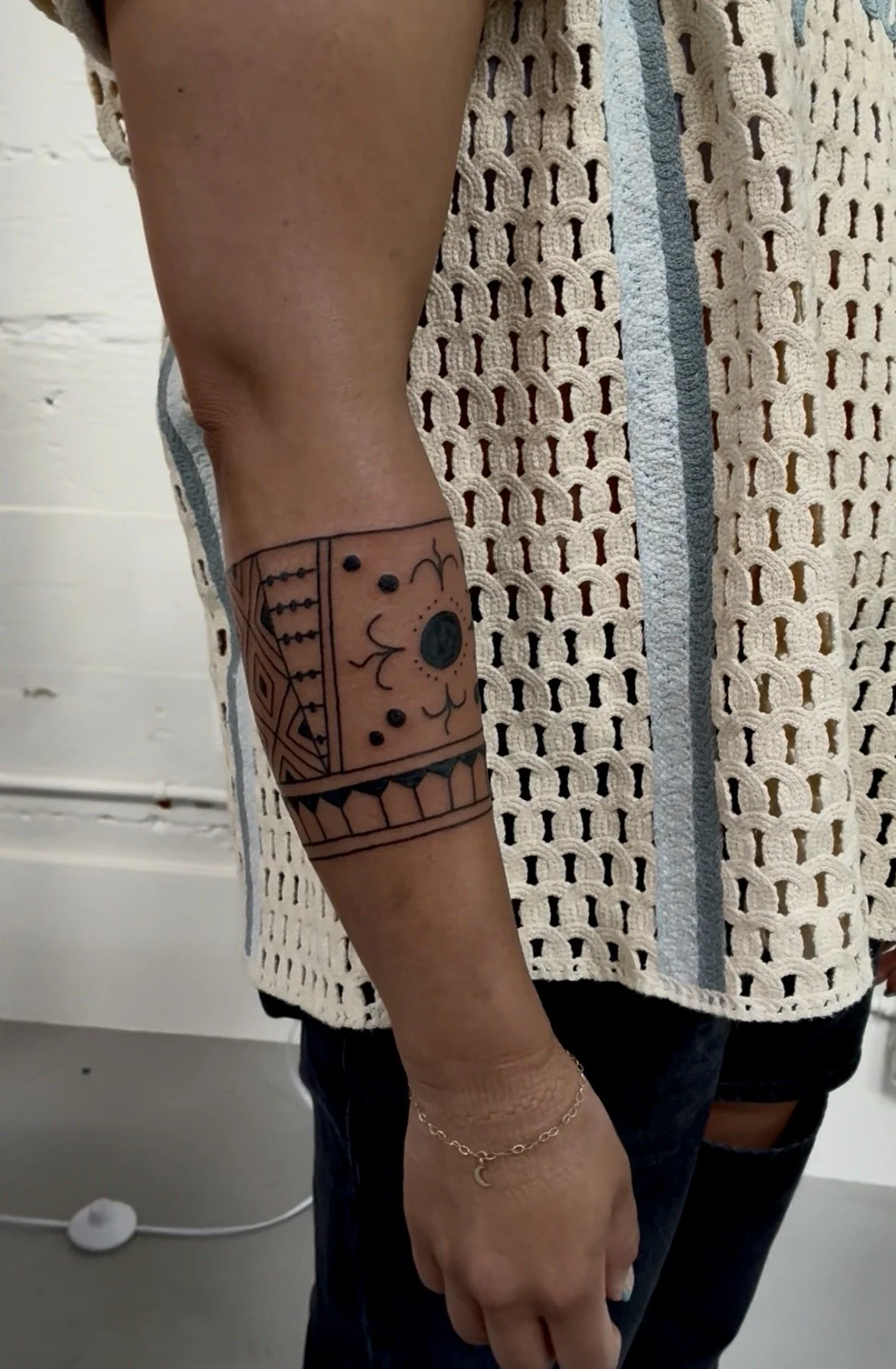 A person's arm with a geometric and floral tattoo, wearing a delicate gold bracelet with a crescent moon charm, standing in front of a crocheted curtain.