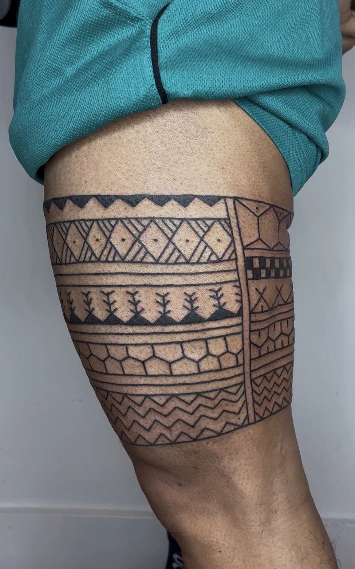 A person's upper thigh covered with a large black tribal tattoo detailed with geometric patterns, lines, and shapes.