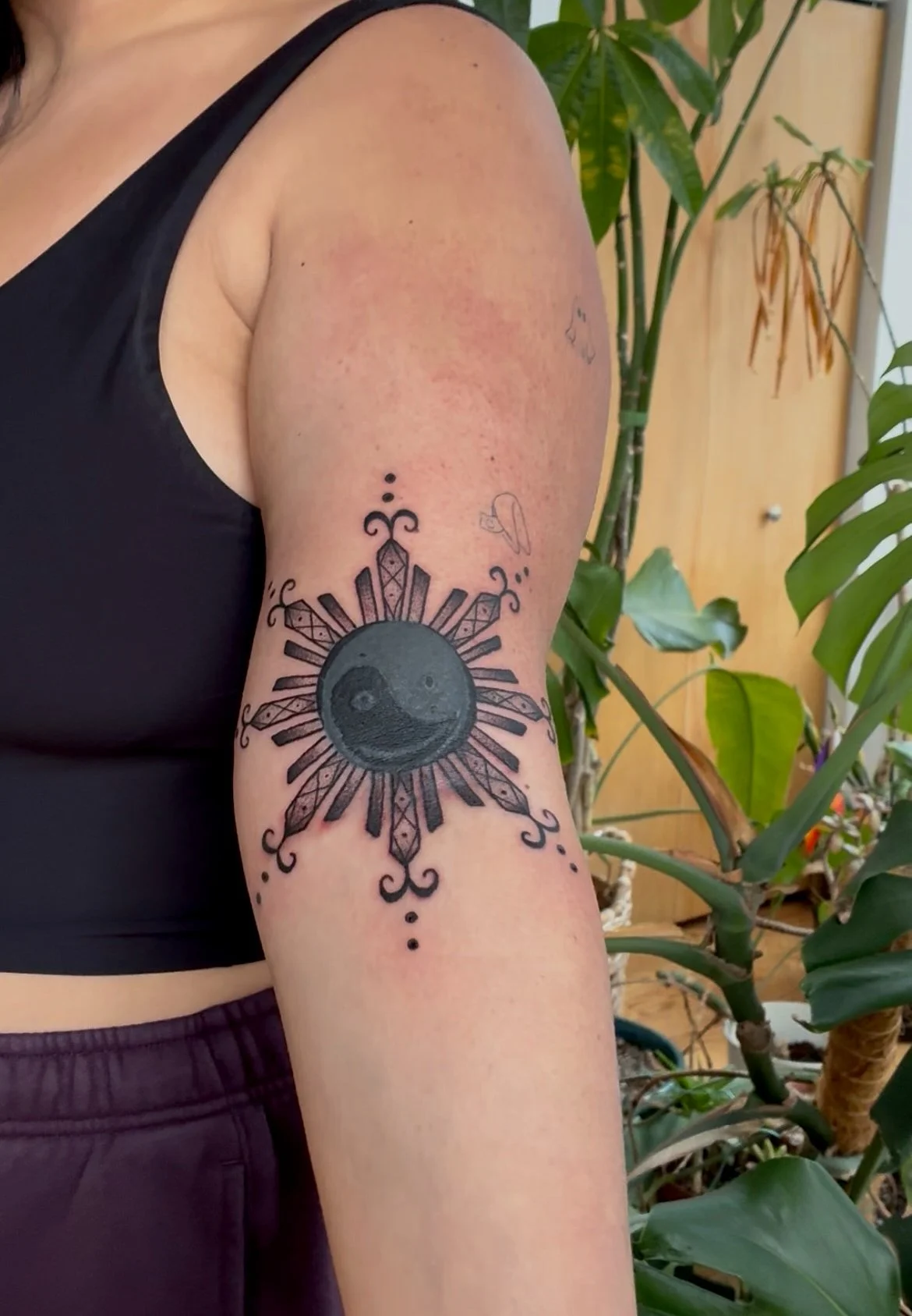 An upper arm tattoo featuring a yin-yang symbol surrounded by sunburst rays and decorative patterns, with a small floral tattoo above the main design.