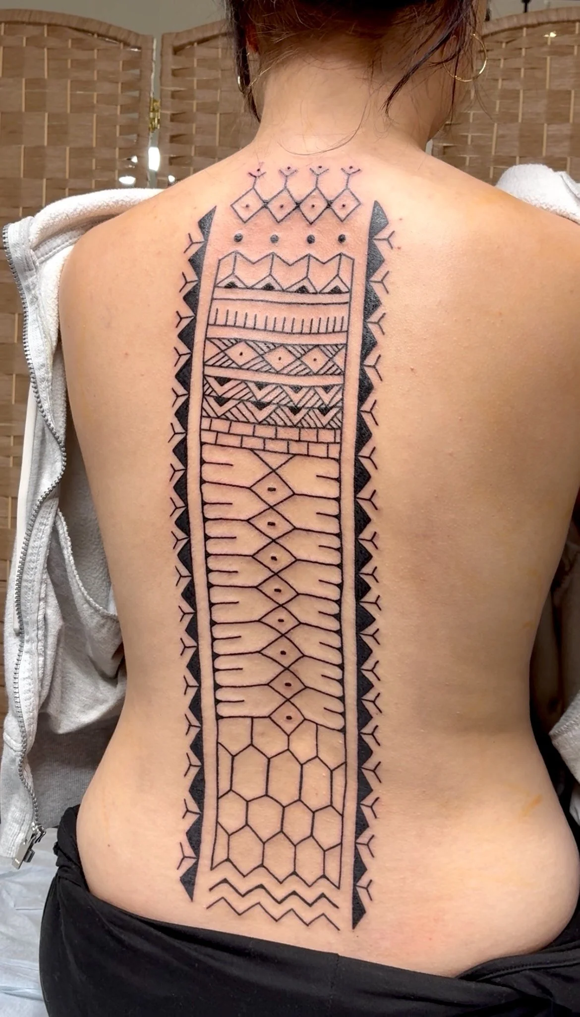 A person's back with a large, intricate geometric tattoo extending from the upper back down to the lower back. The tattoo features patterns such as hexagons, diamonds, lines, and zigzags in bold black ink.