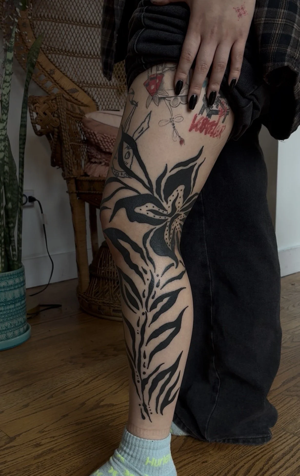 A person with black painted nails showing a large black floral tattoo on their thigh, with smaller tattoos above including the word 'Kobold' in red, and a tattoo of a mushroom with a red cap. The person is wearing dark pants and lime green socks, sta