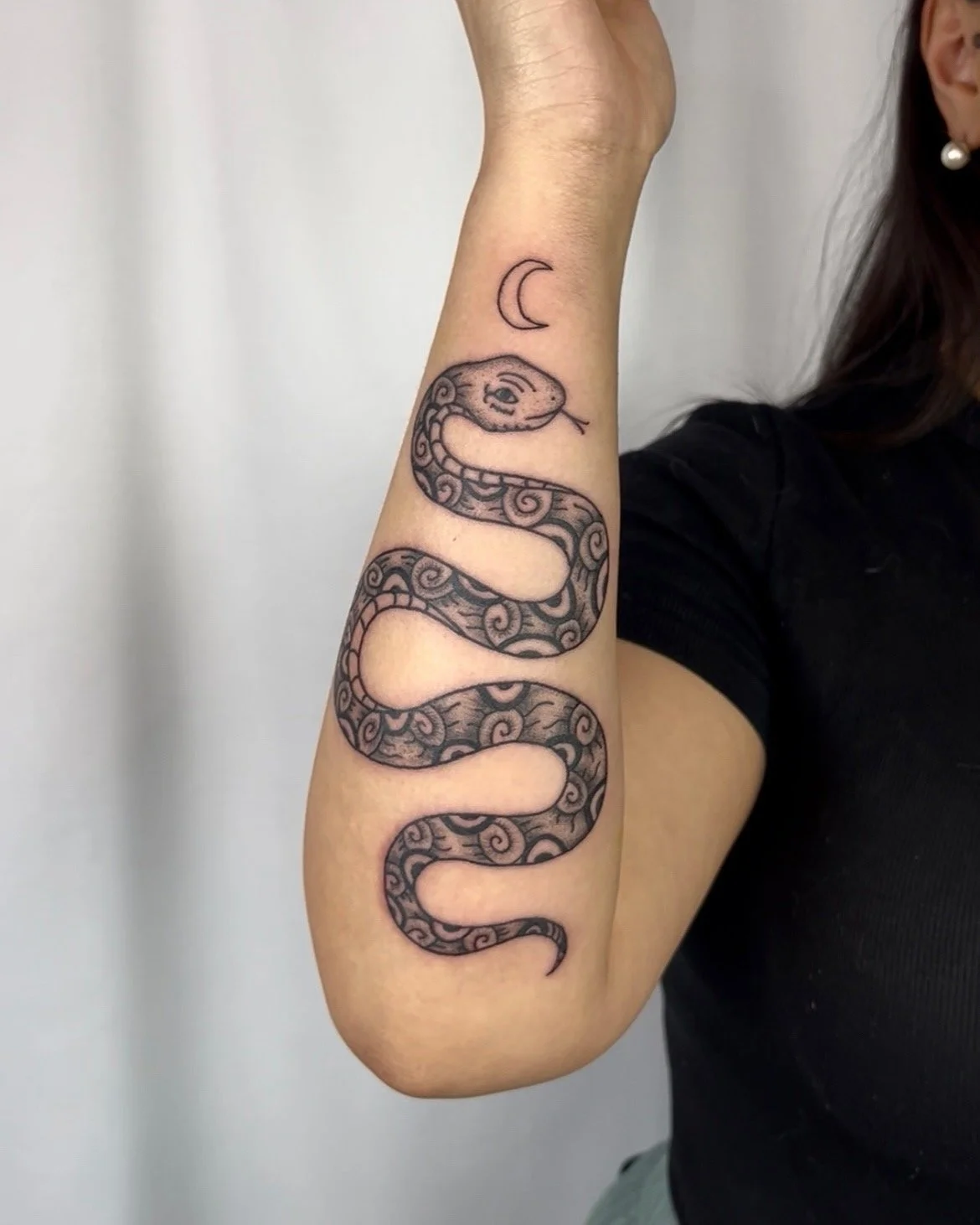 A tattoo of a snake with a detailed pattern and a small crescent moon above it on a person's forearm.