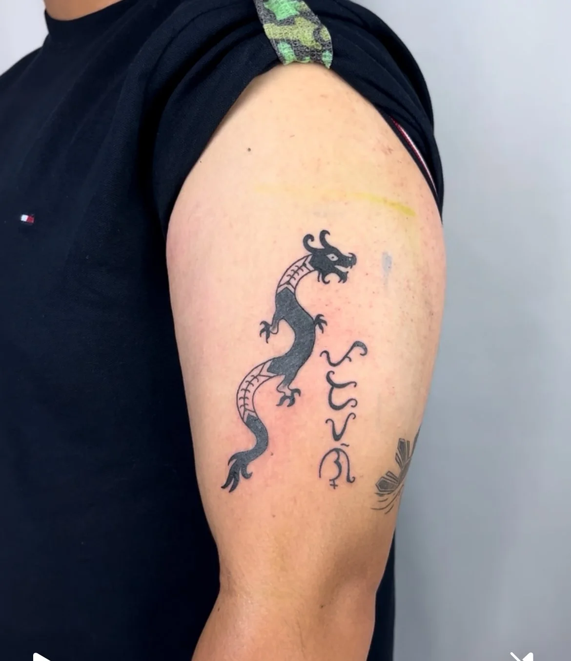 Tattoo of a black dragon on a person's upper arm, with some stylized script or symbols written vertically beside it.