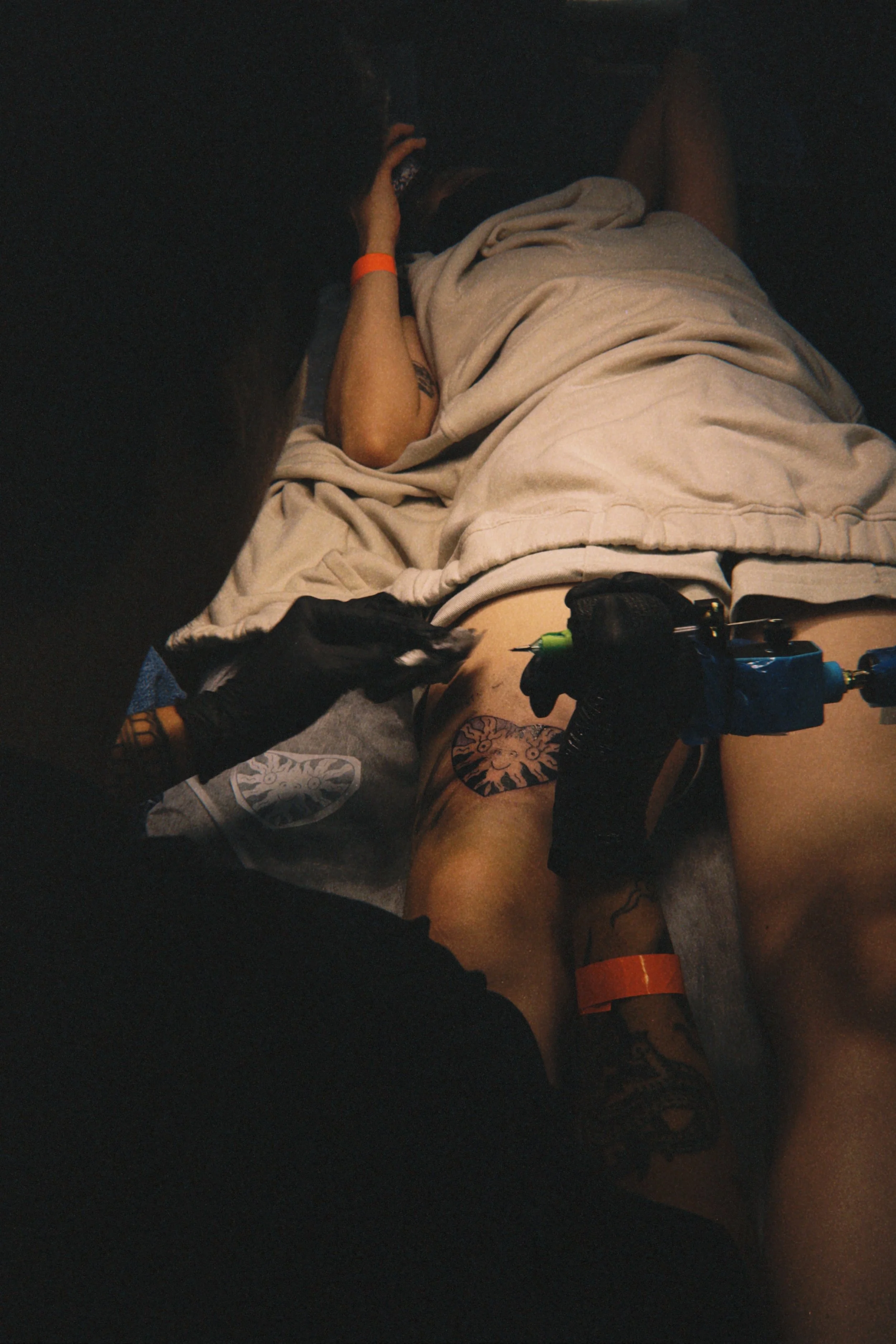 A person lying on a bed with a blanket, tattoos visible on the arm and leg, a wristband on the ankle, and a tattoo on the thigh. The person is using a phone, and a tattoo artist is working on their leg.